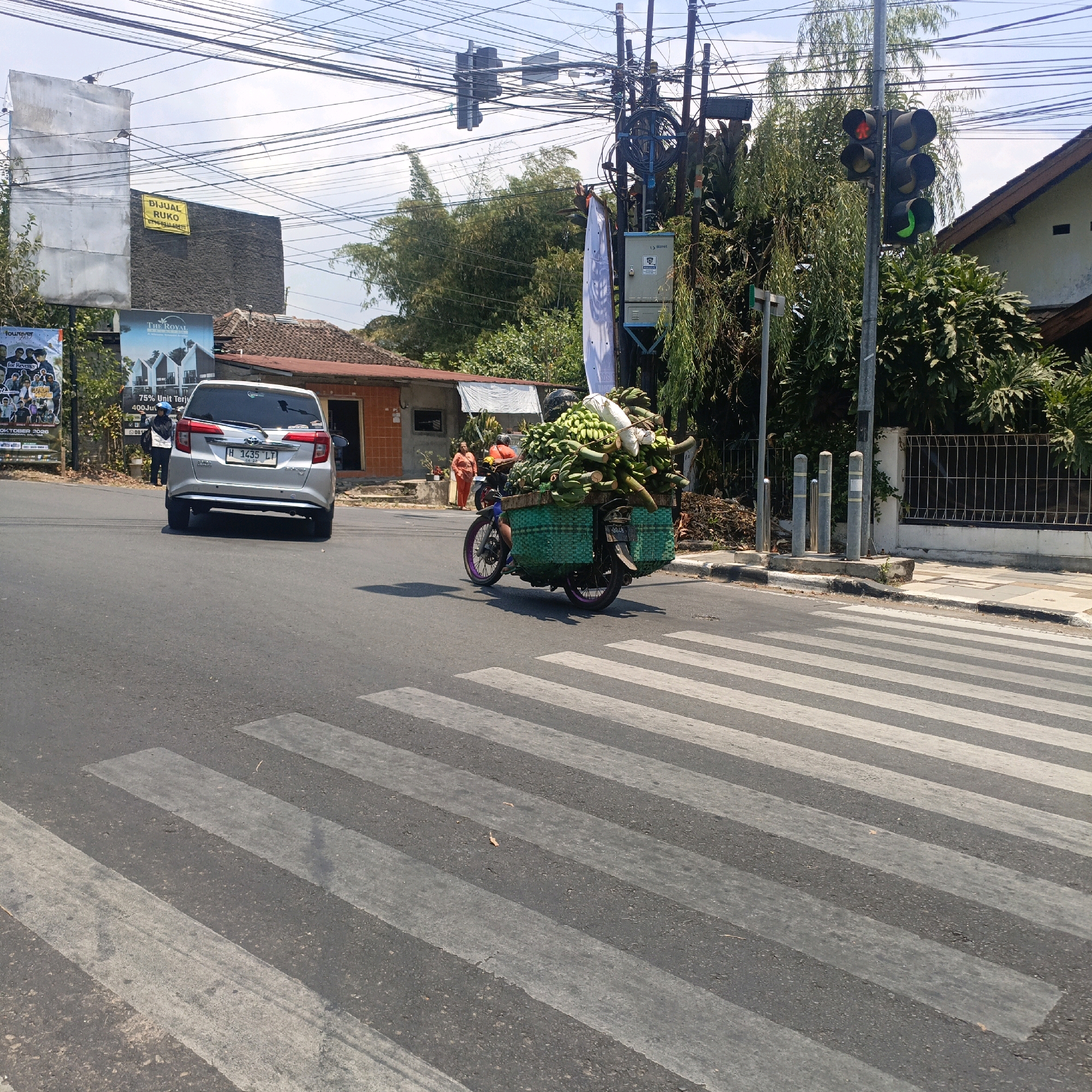 This is a street-level, eye-level, medium shot captured in Salatiga, Indonesia, on a bright, sunny day. The scene is dominated by a busy intersection with a pedestrian crossing in the foreground. A silver Toyota Avanza, with license plate H 1435 LT, is stopped at the crossing, facing away from the camera. To the right of the car, a person on a motorcycle is transporting a large quantity of bananas in green woven baskets attached to the sides of the bike. The motorcycle is also carrying a white sack on top of the bananas. The rider is wearing a helmet.
In the background, a traffic light is visible, with the red light illuminated. Several utility poles with a dense network of wires crisscross the sky. Buildings are present on both sides of the street, some appearing to be commercial establishments with signage, including one that reads "DIJUAL RUKO" (Ruko for Sale). Trees and lush green foliage are scattered throughout the scene, indicating a tropical climate. The pedestrian crossing is clearly marked with white stripes on the asphalt road. The overall impression is a snapshot of everyday life in a bustling Indonesian city.