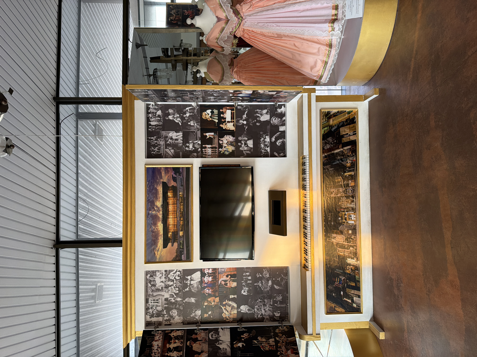 This media file depicts a museum or exhibition display featuring historical photographs and costuming. The primary subjects are a collection of framed black and white photographs arranged in a collage, and a pink, ornate dress displayed on a mannequin. The photographs appear to be historical, showing groups of people in various settings, possibly theatrical or social gatherings from an earlier era. Some images are close-ups of individuals, while others are wider shots of staged scenes. The dress itself is elaborate, with lace detailing and a full skirt, suggesting it is a historical costume.
The scene is set indoors, with a neutral wall and a polished, reflective surface behind the mannequin and dress, which mirrors the display. A flat-screen television is mounted on the wall below the photographs. To the right of the television, a decorative element resembling piano keys is visible. Another framed artwork, a vertical landscape with warm, sunset lighting, is displayed to the right of the photographs. The overall impression is one of a curated exhibition intended to showcase historical artifacts and imagery. There are no clear cues about the time of day or weather, and the emotions conveyed are neutral, focused on the historical presentation.
The location context provided is Odesa, Ukraine. While there are no directly visible text elements or explicit location cues within the image itself, the exhibition is presumed to be related to the history of Odesa.