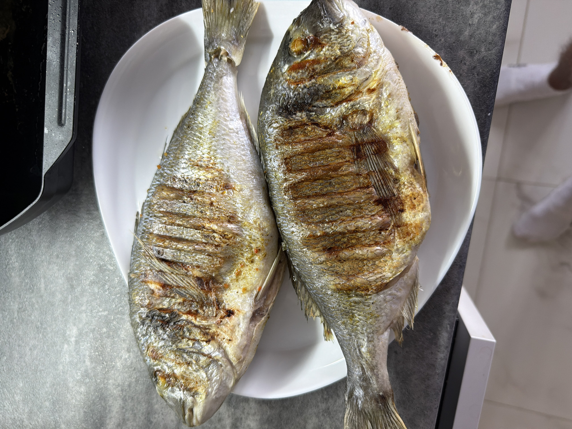 A top-down shot shows two cooked whole fish, likely gilt-head bream (Sparus aurata), arranged on a white plate. The fish are positioned side-by-side. The fish have been cooked in a way that leaves noticeable grill marks on their bodies. The fish are intact, with heads, tails, and fins present. The plate rests on a counter surface, with a portion of what appears to be a black appliance visible on the left side. There's a glimpse of white flooring and part of a person's lower leg in white socks on the right side. The overall lighting is bright. There is no specific visual evidence in the image to indicate the location is Chaikovsky, Russia.