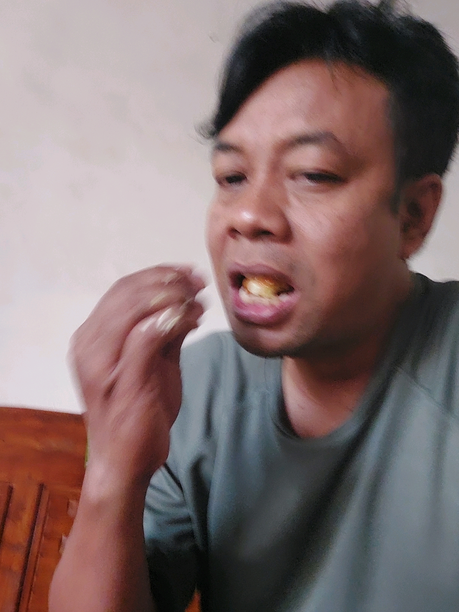 In Sarangan, Indonesia, a close-up shot shows a person with dark hair and medium-toned skin. The individual is in the process of eating, with food visible in their open mouth. Their right hand is near their mouth, and it appears to be holding or bringing food to their mouth. The person is wearing a plain, dark-colored t-shirt. The background is a plain wall. A wooden furniture piece is partially visible in the lower left corner, suggesting the setting might be indoors. The lighting is soft and diffused, casting a gentle glow on the subject. The image is taken from a slightly high angle, looking down at the person.