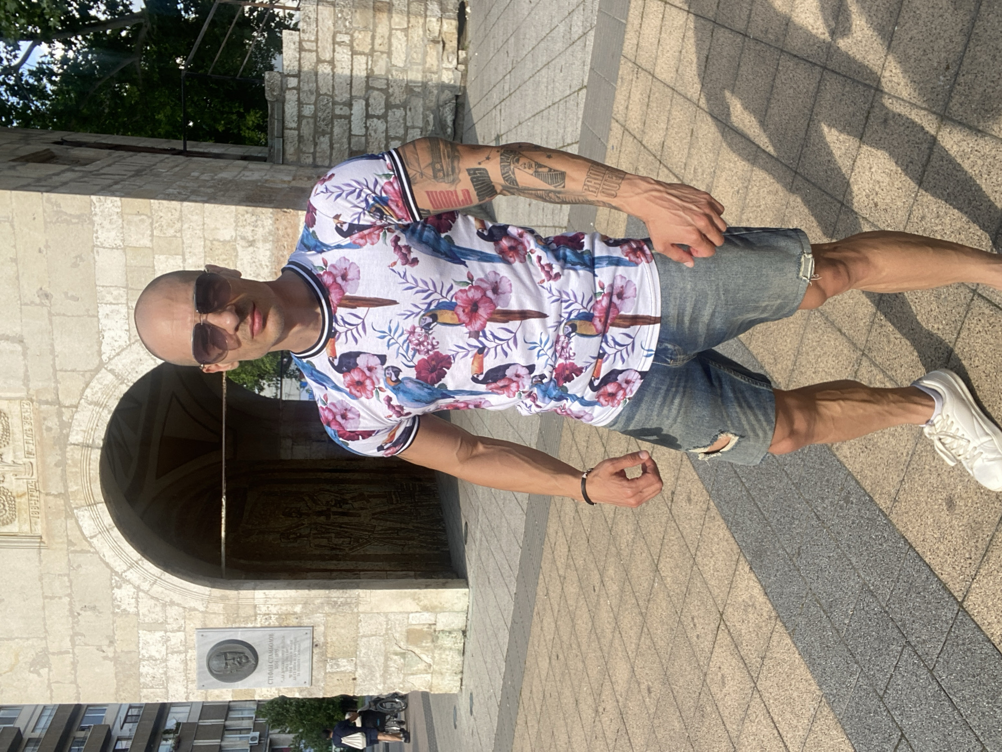 An adult male stands centrally in an outdoor urban setting in Pleven, Bulgaria, in front of a historic stone building. The man is bald and wears dark aviator-style sunglasses. He is dressed in a white short-sleeved t-shirt patterned with blue parrots, toucans, and pink and purple flowers, distressed blue denim shorts, and white low-top sneakers. His physique is muscular, with visible veins on his forearms and calves. His left forearm is covered in tattoos, including a depiction of a pharaoh or sphinx head, a car, and text. A plain black bracelet adorns his right wrist. He faces the camera with a neutral expression.
Behind the man is a large, light-colored stone building with a prominent arched entrance. Above the arch, an inscription reads "1880-1904 Г. Н. БАНК" (1880-1904 G. N. BANK). To the left of the arch, a rectangular plaque with a relief bust is affixed to the wall. Text on the plaque identifies the individual as "СТЕФАН СТАМБОЛОВ 1854-1895 ПРЕМИЕР-МИНИСТЪР НА БЪЛГАРИЯ" (Stefan Stambolov 1854-1895 Prime Minister of Bulgaria). Through the archway, a decorative wooden or metal door with relief work is visible.
The ground is paved with rectangular tiles in alternating light and dark grey shades. Strong sunlight casts distinct shadows on the pavement. In the far left background, a multi-story modern residential building is partially visible, and two distant figures appear, one pushing a stroller or wheelchair. Green foliage and trees are visible to the right of the stone building.
