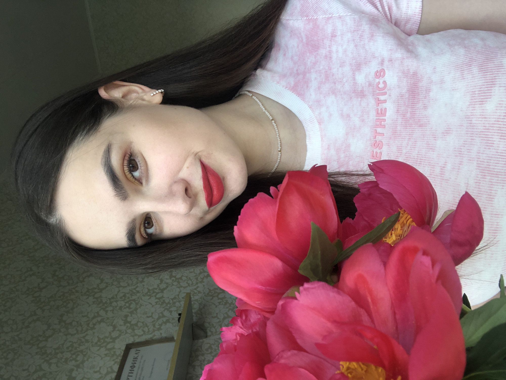 A young woman with long, dark hair and brown eyes is holding a bouquet of bright pink peonies. She is wearing a white t-shirt with a pink tie-dye pattern and the word "ESTHETICS" printed on it in pink. She also has on a delicate pearl and gold beaded necklace and small hoop earrings. Her lips are painted a bold red. She is smiling slightly. In the background, there is a patterned wallpaper and a framed certificate on a shelf. The image appears to be taken indoors, possibly in a home in Noviy, Russia.