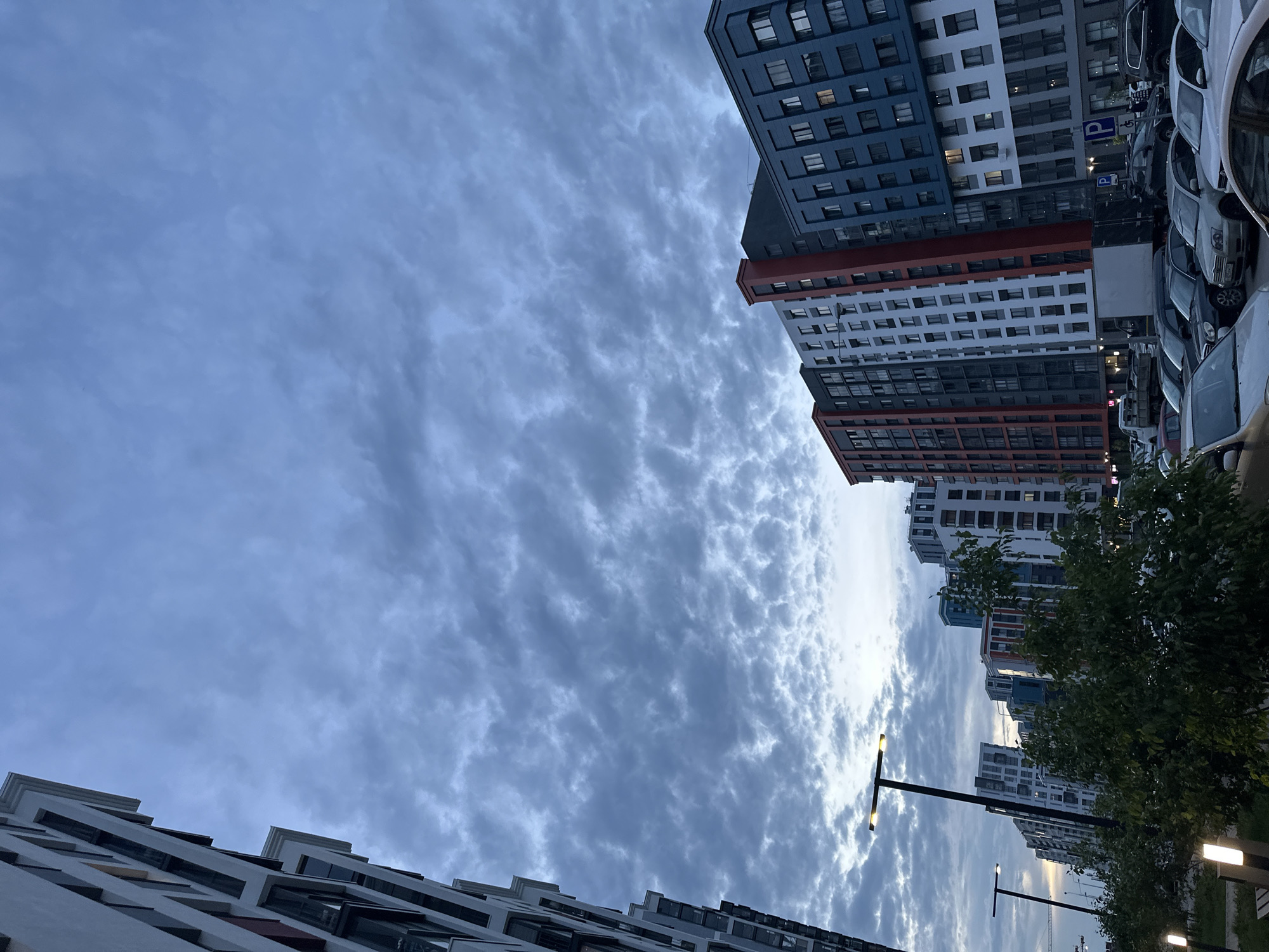 The image depicts an upward-angled view of a modern residential area under a cloudy sky, potentially located in Perm, Russia. Several high-rise apartment buildings are visible, featuring various architectural designs and color schemes, including white, blue, gray, and red accents. Cars are parked alongside one of the buildings. Streetlights illuminate the area. Tree foliage is present near the right side of the frame. The sky is filled with altocumulus clouds.