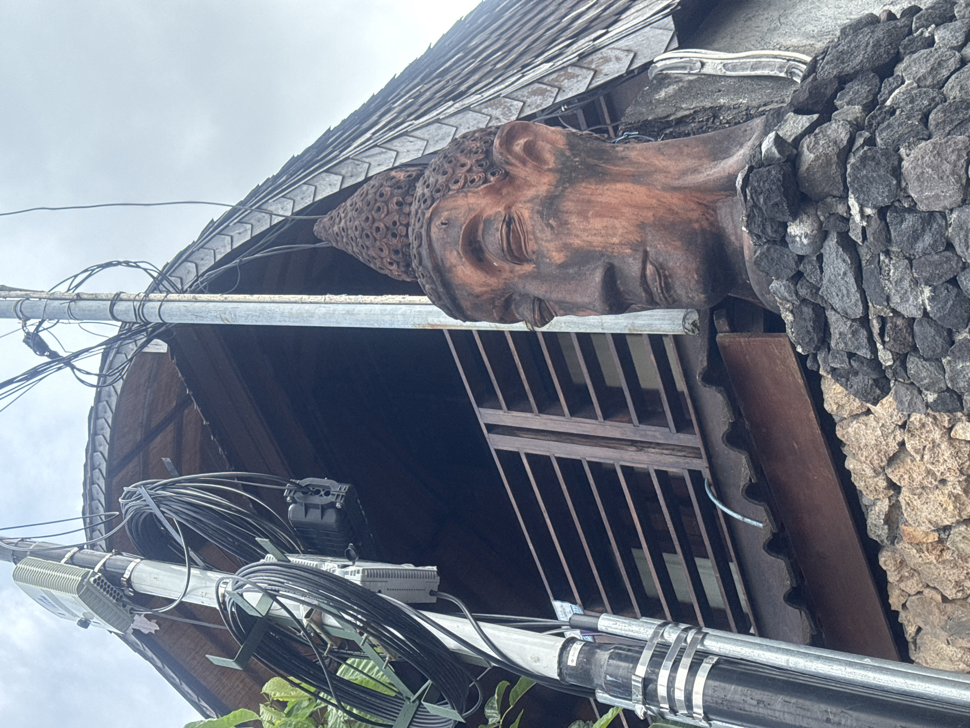 The image captures a close-up view of a building detail in North Kuta, Indonesia. A statue of a serene Buddha head, crafted in a dark, possibly wooden, material, protrudes from a structure built with dark gray volcanic rocks. The building also features a tiled roof and a dark brown metal grate. A complex network of black and gray cables and wires is attached to a galvanized metal pole running horizontally across the frame. In the background, the sky is overcast. The overall perspective suggests the photo was taken from a low angle, looking up at the building structure.