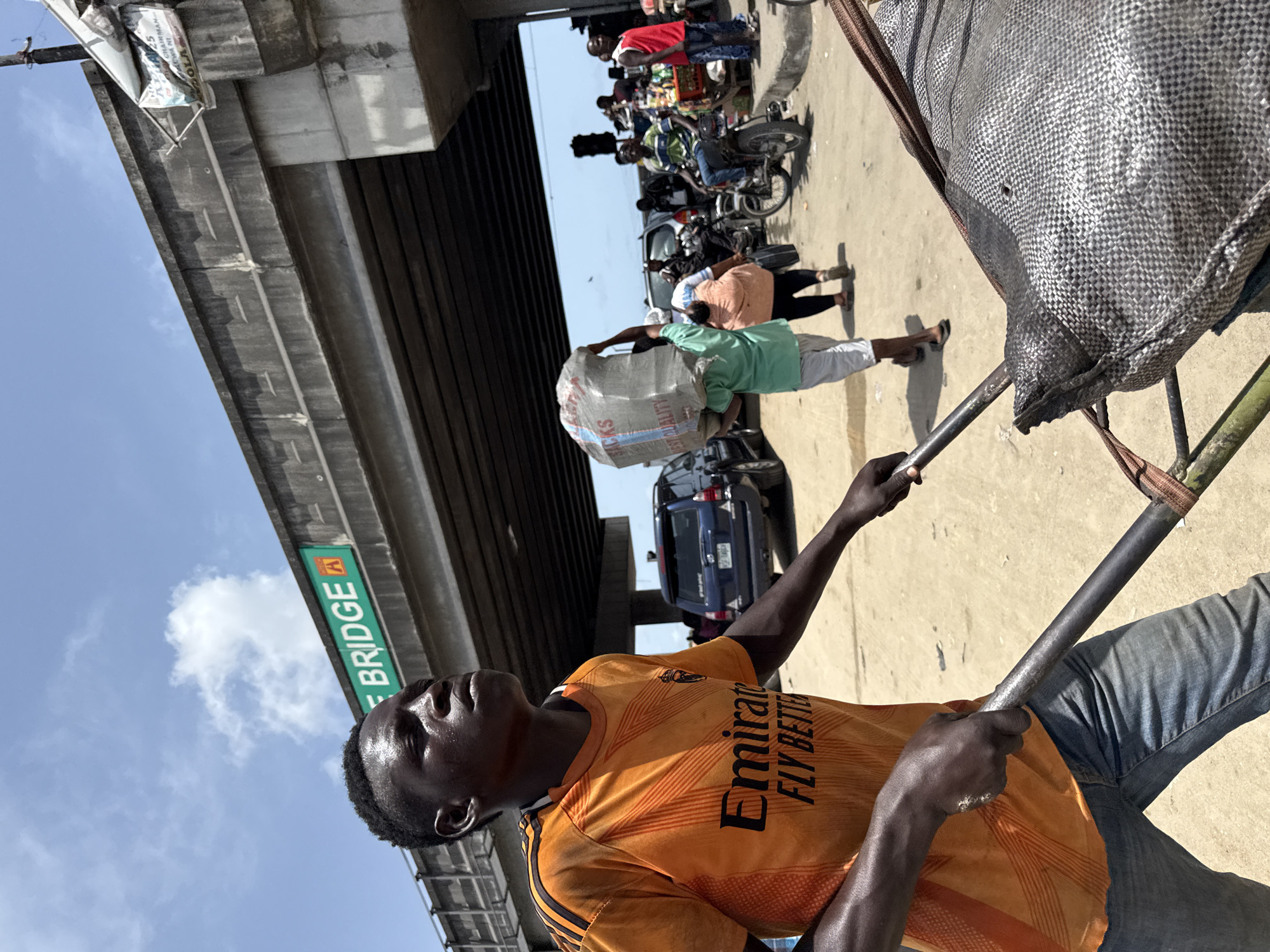 A street-level perspective shot of a man pulling a cart under a bridge in Lekki, Nigeria. The man, wearing an orange soccer jersey with black stripes and blue jeans, holds the cart handles. A large silver-gray bag sits on the cart. In the background, under the bridge, are people, motorcycles, and a blue car. A green sign labeled "LEKKI BRIDGE A" is visible. The sky is blue with a few clouds. Another person is also seen carrying a bag on their head.