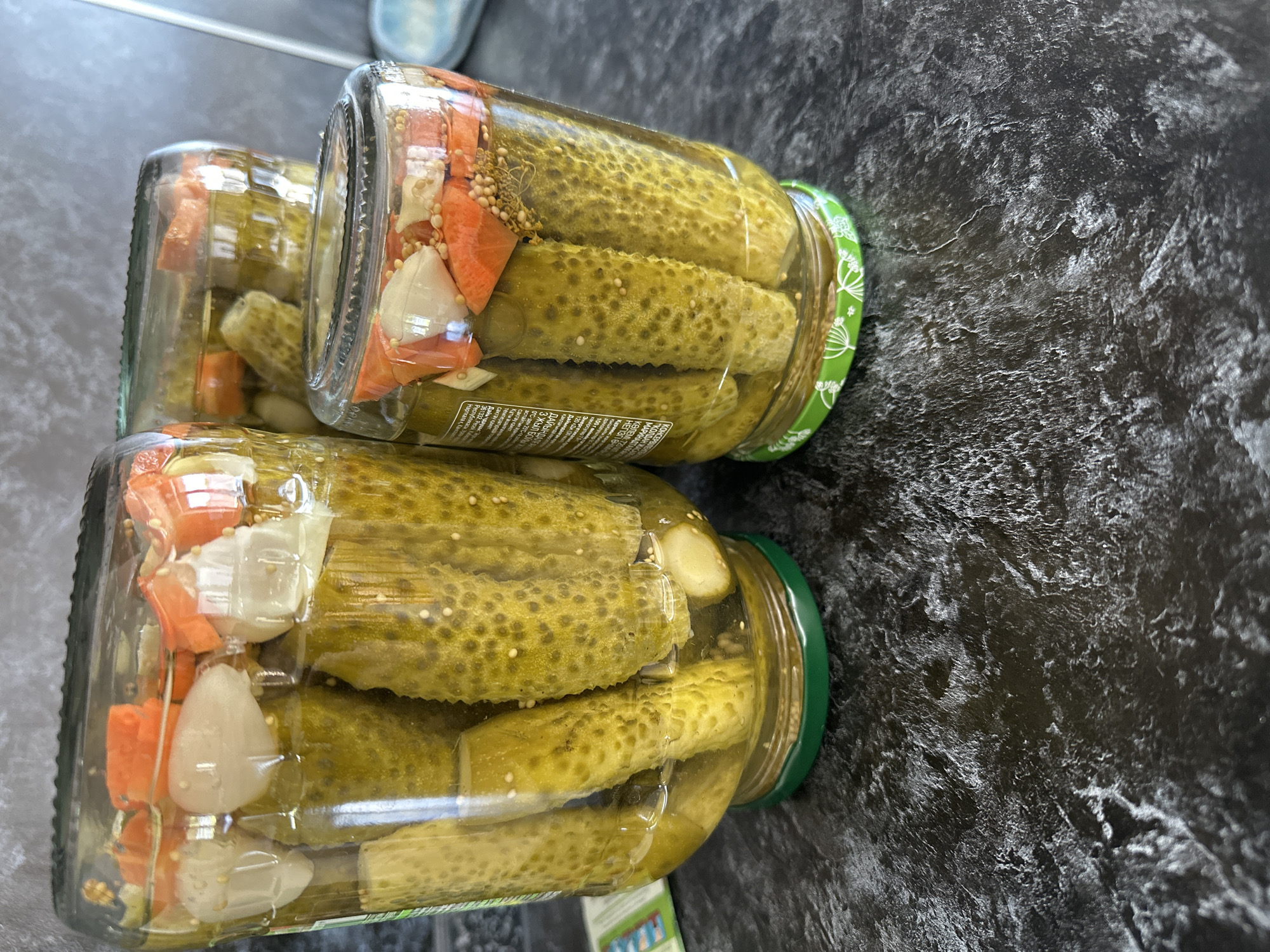 The image shows three glass jars of pickles on a dark, textured countertop. The jars are filled with whole pickles, brine, and visible pieces of chopped carrots and garlic. The jar in the foreground is the smallest and is slightly to the right of center. Behind it, to the left, is a taller, wider jar. A third jar is visible in the background, partially obscured by the other two. The jar lids appear to be metal, with the foreground jar having a green lid with a white floral pattern. The countertop has a marbled or stone-like appearance with lighter gray and white veining against a dark background. There are no people or animals present in the image, and no visible indication of the location being in Белозерки, Russia. The scene is static and focuses on the preserved food items.