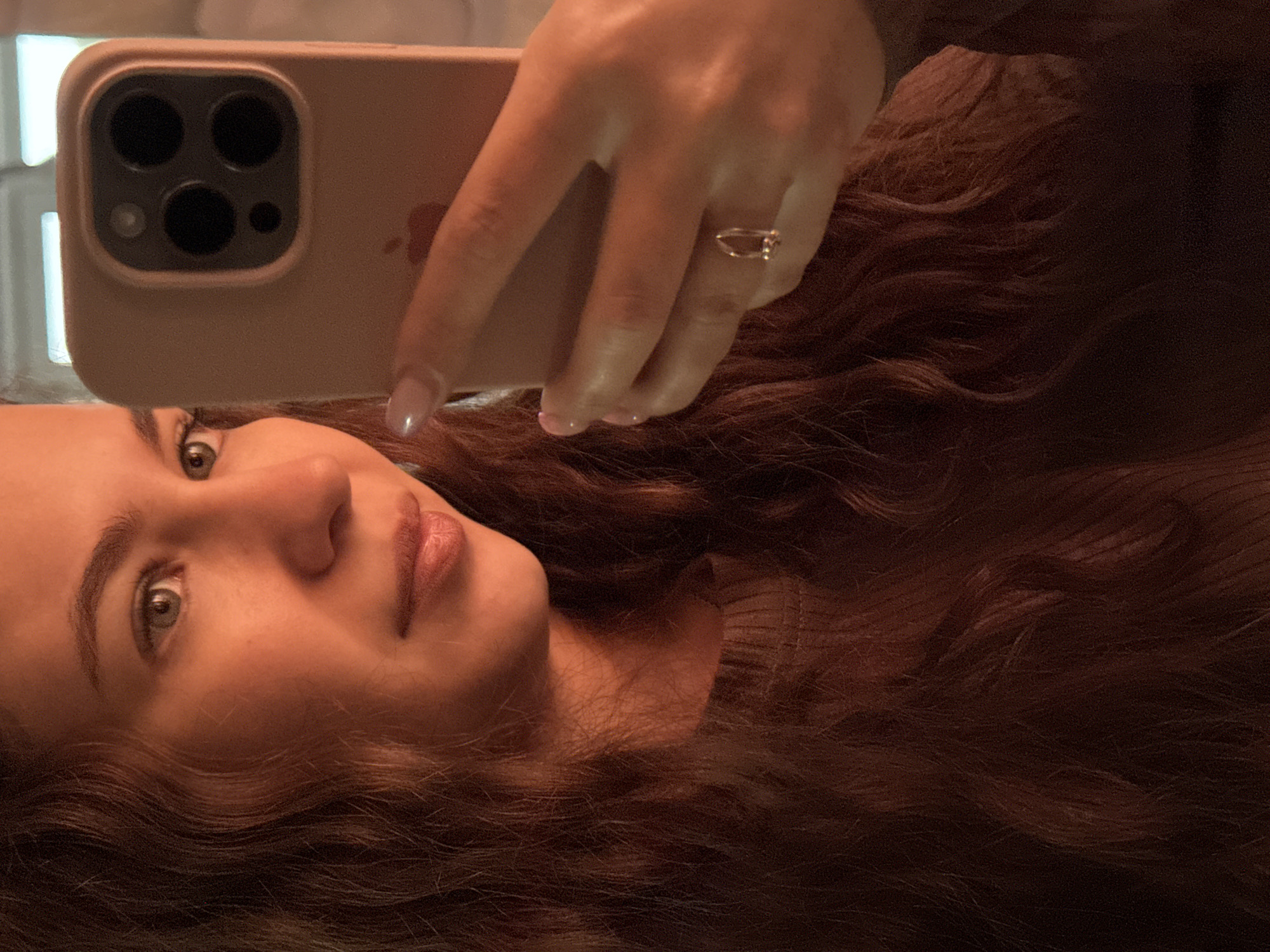 A woman is taking a selfie in a mirror with a smartphone. She has long, wavy brown hair and is wearing a ring on her left hand. The smartphone has a beige case with multiple camera lenses. The background is out of focus, showing a window with blurry light. The location is unspecified, as the city name provided is not visually discernible from the image.