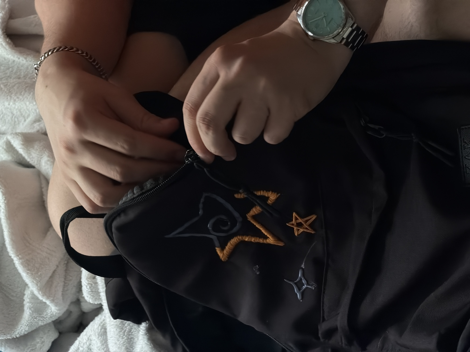 A person wearing a silver watch and bracelet is zipping up a black backpack with embroidered star designs. The surrounding environment includes a white towel or blanket. The location is not discernible from the image.