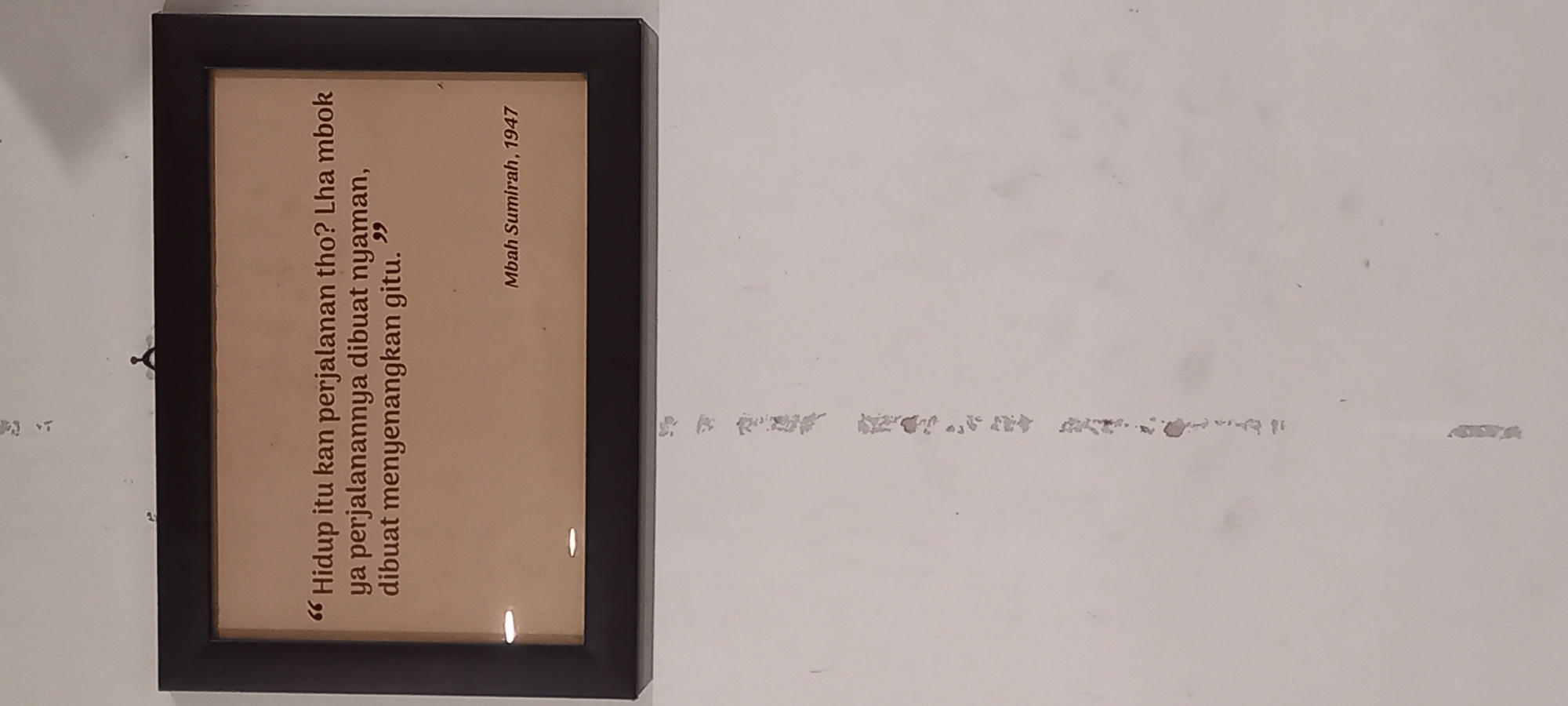 The image displays a framed quote hanging on a white wall. The quote, written in a dark font on a light brown background, reads: "Hidup itu kan perjalanan tho? Lha mbok ya perjalanannya dibuat nyaman, dibuat menyenangkan gitu." Below the quote, it is signed "Mbah Sumirah, 1947". The frame is dark and appears to be made of wood. There are some faint, vertical marks of peeling paint or dirt on the white wall below the frame. The image appears to have been taken in an indoor setting, as there is no visible indication of an outdoor environment. There is no information available to determine the specific location in the city of Timbau, Indonesia.