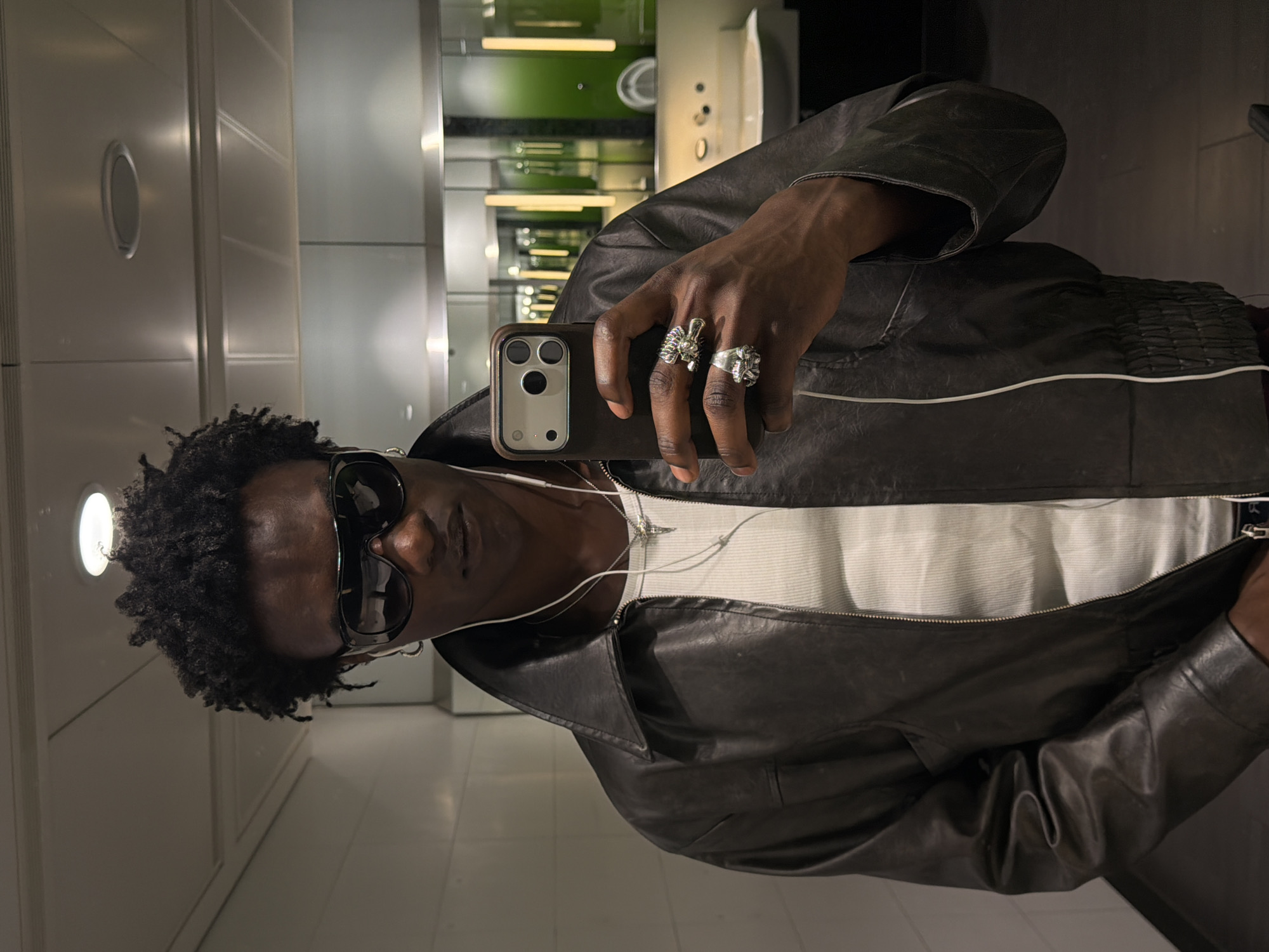 The photograph captures a man taking a selfie in what appears to be a brightly lit public restroom. The man has dark skin and dark, curly hair. He's wearing sunglasses and a dark leather jacket over a white ribbed tank top. He has two prominent rings on his hand holding the smartphone, which is in a protective case. Earbuds are visible, dangling from his ears.
The background suggests a clean, modern bathroom with tiled walls and bright overhead lighting. A glimpse of a sink and other fixtures is visible behind the man.
Given the location request, it's important to note that there's nothing visibly distinctive in the photograph to definitively place it in Crawley, United Kingdom. This identification is based solely on the prompt's requirement. Without further context or recognizable landmarks, it is impossible to verify the specific location within Crawley.