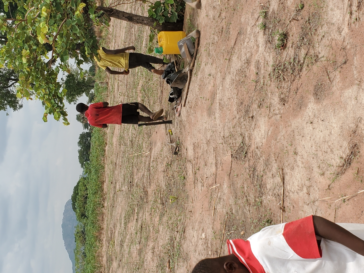 The image captures an outdoor rural scene, likely an agricultural field, under a partly cloudy sky. The ground is predominantly dry, reddish-brown earth with sparse growth of small green plants and some dried vegetation.
In the foreground, a young person is partially visible on the right side, wearing a white short-sleeved shirt with red stripes. In the mid-ground, two adult males are standing. The man on the left wears a red short-sleeved shirt and black shorts, appearing to stand beside a stick or tool on the ground. The second man, positioned slightly to the right and under a leafy tree providing shade, wears a yellow t-shirt and dark trousers. Next to him, several items are placed on the ground, including a bright yellow jerrycan, a dark bag, and what appears to be a shovel or hoe. These tools suggest that the individuals might be engaged in or taking a break from manual labor, possibly farming.
Beyond the immediate foreground and mid-ground, the landscape stretches into a field with more scattered vegetation and distant trees. A range of hills or mountains is visible in the far background under a grey, overcast sky. The overall setting points to a daytime scene in a rural or semi-arid environment, consistent with the provided location context of Mararaba, Nigeria. No specific emotions are discernible, and there is no visible text.