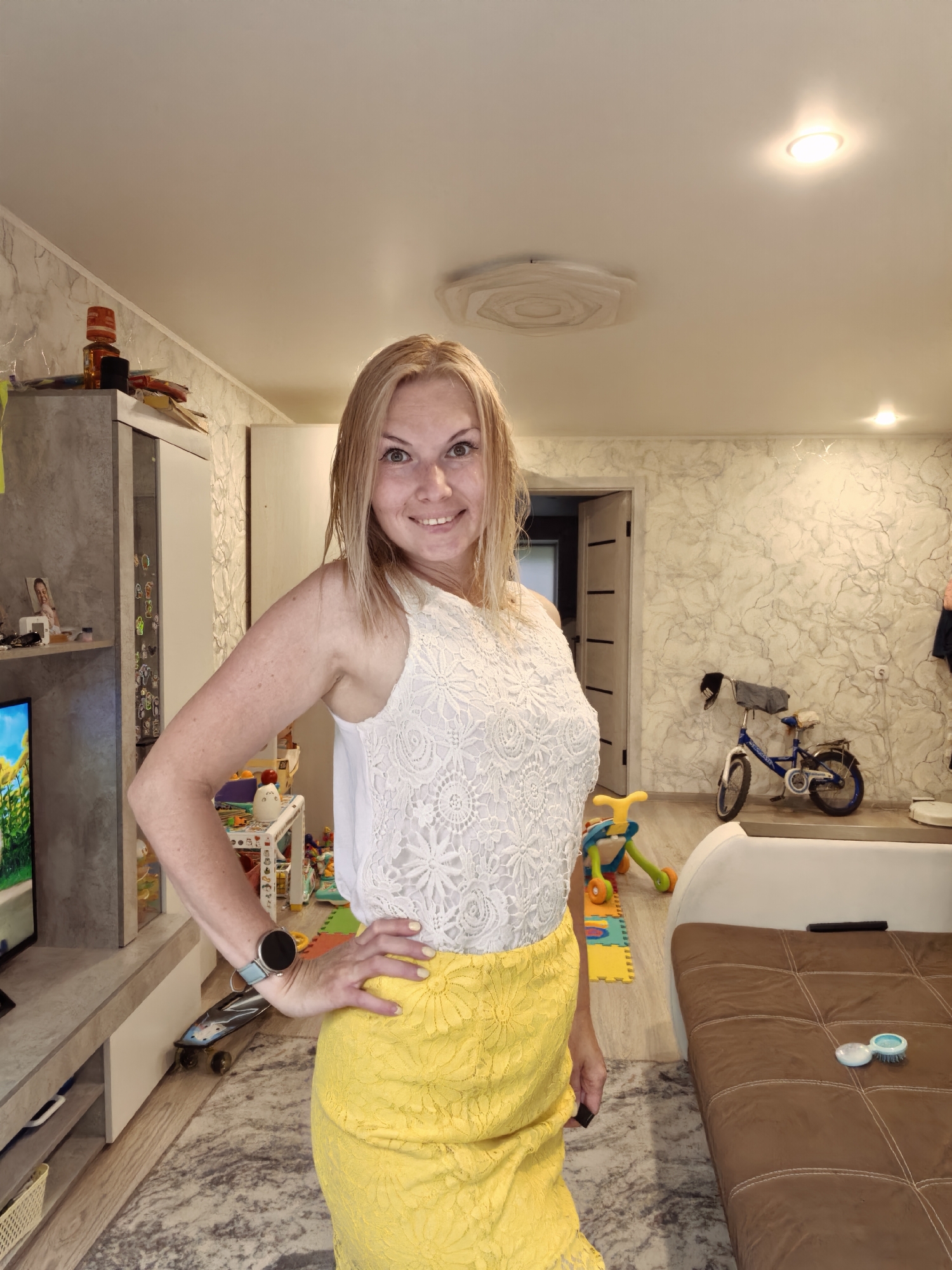 A blonde woman in a white lace top and a yellow skirt stands in a living room, posing for a photo. The room has a patterned wallpaper, a TV on a stand, a couch, children's toys, and a bicycle. There are also lights on the ceiling. The woman is smiling and has her hand on her hip. The image does not contain enough information to determine the location of the city of Sterlitamak, Russia.