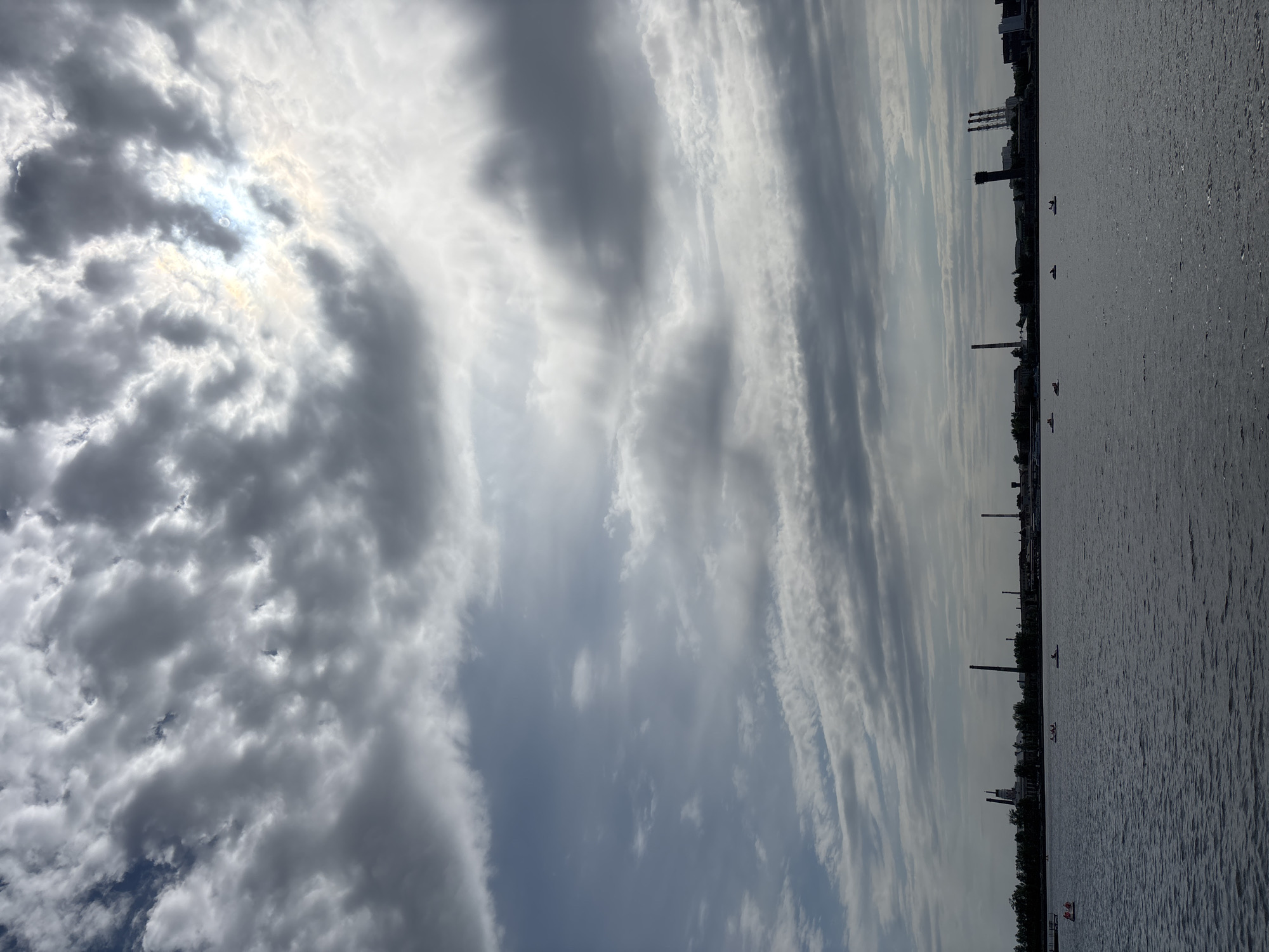 The image depicts a body of water adjacent to a city skyline, with a dramatic sky overhead. The sky is filled with an array of cloud formations, ranging from thick, billowing clouds in the foreground, potentially obscuring the sun, to elongated, linear clouds stretching across the sky. The water surface appears calm, with subtle ripples reflecting the light. Along the shoreline, there are industrial structures such as chimneys or towers and what appear to be buildings. Small boats are scattered on the water, some of them carrying people. The scene conveys a sense of scale, with the vastness of the sky contrasting with the industrial presence along the water's edge.