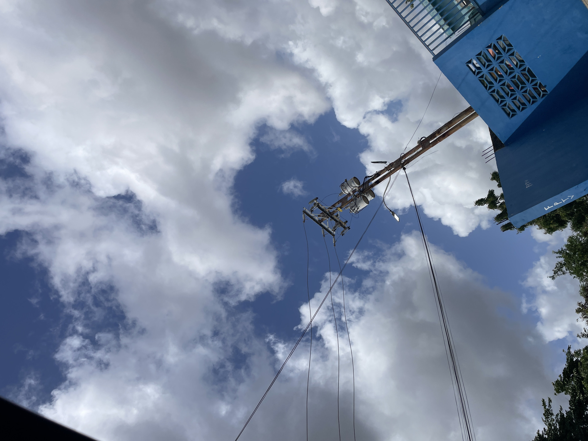 The image captures a view looking upwards towards a blue sky filled with white, puffy clouds. A wooden utility pole, weathered and leaning slightly, stands prominently in the center-right of the frame. It is equipped with electrical transformers and wires that extend across the sky. In the lower right quadrant, a bright blue, angular structure with a decorative vent is visible, suggesting a building or wall. To the left and bottom, the tops of lush green trees can be seen. The scene appears to be an outdoor setting in a city, likely Juan Griego, Venezuela, given the context. There are no people or animals visible in the image, and no discernible actions or interactions are taking place. The lighting suggests it is daytime, with the sun illuminating the clouds and the scene.