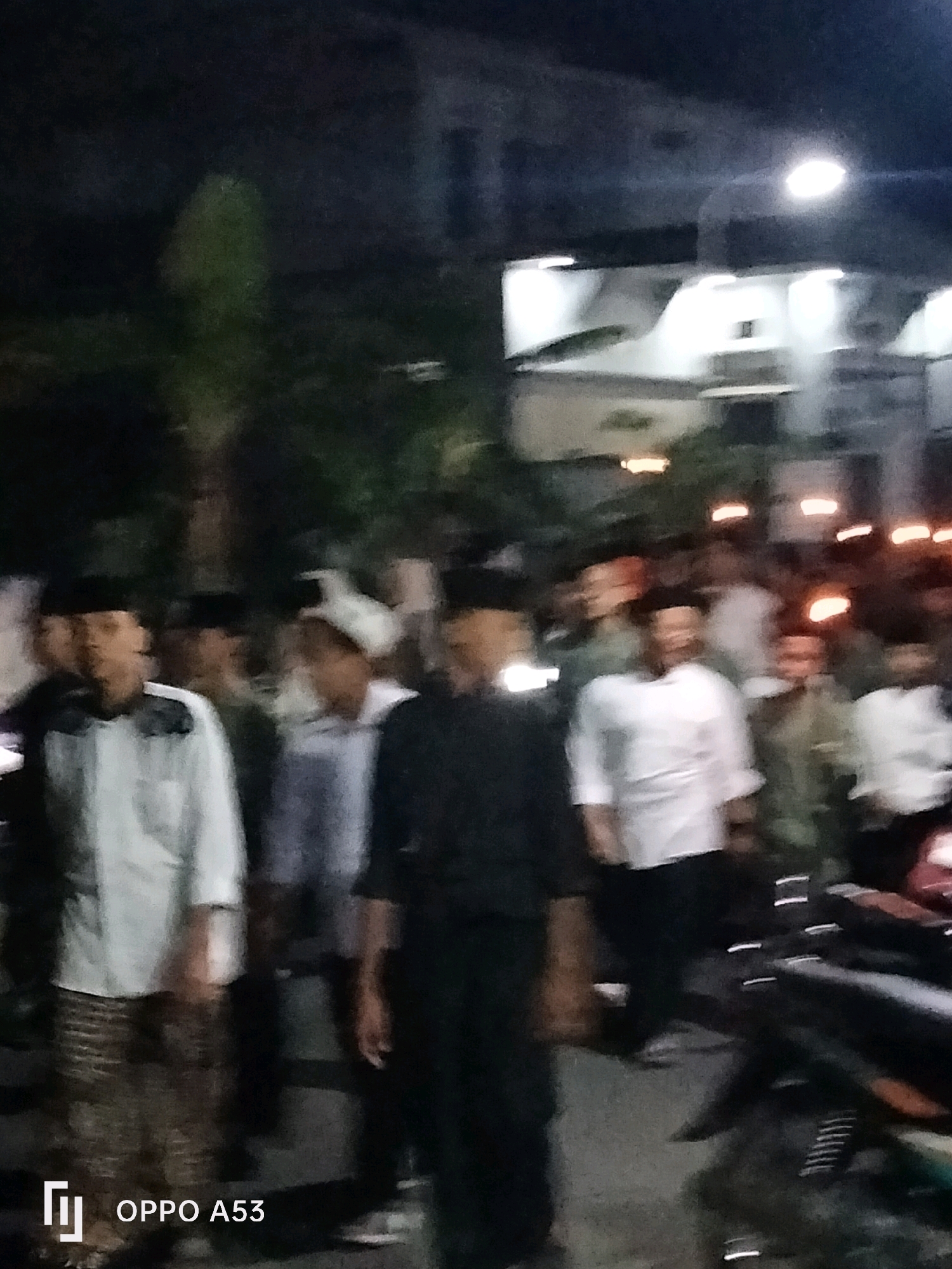 In Bendorejo, Indonesia, a group of individuals are gathered outdoors at night. The scene is dimly lit, with several visible light sources including streetlights and torch-like flames held by some people. A building with illuminated windows is present in the background. The people, dressed in what appears to be traditional or religious attire, are blurred due to motion and low light conditions. One individual in the foreground is wearing a white long-sleeved shirt and patterned pants. Another person is wearing a dark shirt and pants. The overall atmosphere suggests a public gathering or procession.
