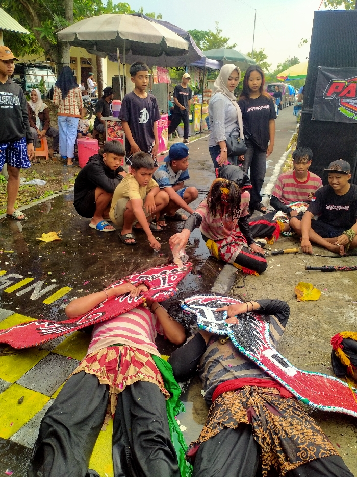 An outdoor scene in Jenggrik, Indonesia, depicts a cultural event on a wet, paved surface. In the foreground, two individuals lie prone on the ground, dressed in dark trousers, patterned loincloth-style skirts, and striped shirts (one red/white, one grey/black). Their faces are obscured by large, elaborate, wing-shaped headpieces (one red/white, one black/white/red). A third individual in ceremonial attire, with a masked face or heavy face paint and disheveled hair, kneels near their heads, pouring liquid from a clear plastic bottle onto the head of one prone figure.
Numerous spectators observe the scene. A group of approximately seven young males are positioned around the central figures, some squatting, some sitting, some standing. Two young females stand observing on the right. Additional individuals and market stalls with umbrellas are visible in the background. The paved ground features yellow and black painted stripes and is wet with puddles reflecting the overcast sky. Large black speaker boxes, one with a partial "PA***" logo, are present on the right. Vegetation and a light-colored building are visible in the background.