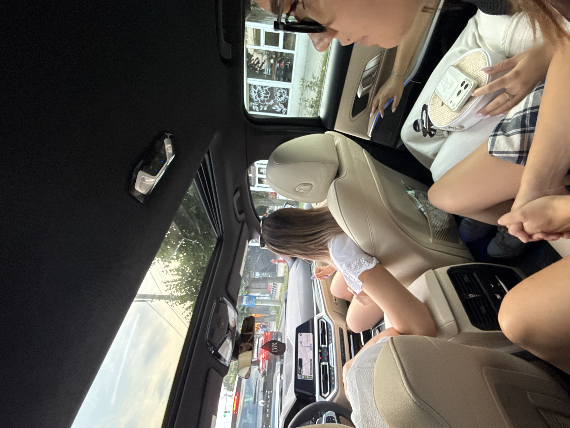 The image shows the interior of a car, with a panoramic sunroof open, revealing a sky with contrails and trees. Inside the car, two women are visible in the back seat. One woman, wearing a white top, is seated in the front passenger seat, looking forward. Her legs are crossed, and she is holding a phone. The second woman, wearing glasses and a black top, is in the back seat behind the driver, looking to her right. She has a white bag with a phone on top resting on her lap. The car's dashboard and navigation screen are visible in the foreground. Outside the car, through the windows and sunroof, a street scene with buildings and traffic can be seen. The license plate of a car in front reads "B967 ESP". The setting appears to be a city street in Onești, Romania, given the presence of what appears to be a street sign with "Onesti" visible in the background.