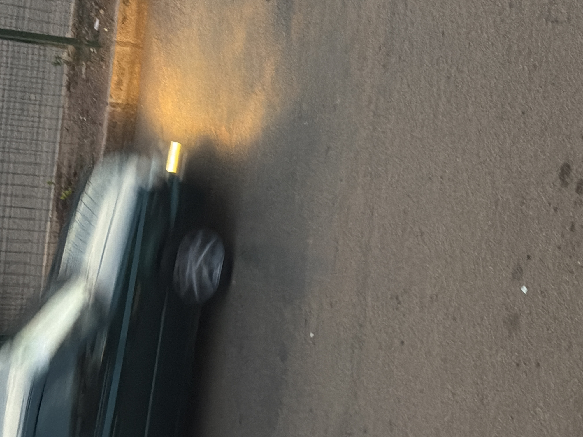 A dark green car is captured in motion on a paved road in Bukuru, Nigeria. The car's headlight illuminates the road ahead with a bright yellow glow, casting a warm light on the asphalt and a yellow curb. The motion blur indicates the vehicle is moving at a moderate speed. Behind the car, a wire fence separates the road from a dirt embankment. The overall scene suggests an urban or suburban setting in the late afternoon or early evening, given the warm lighting.