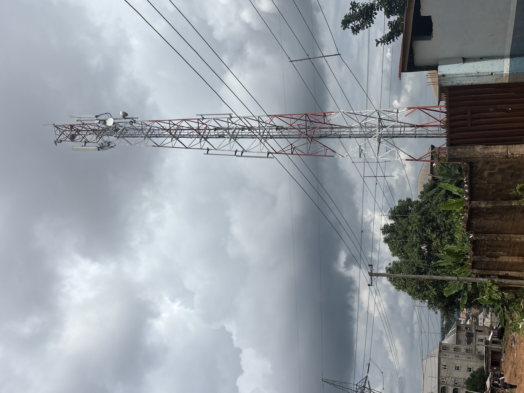 The image captures a cell phone tower situated in Nkpor, Nigeria. The tower, a prominent metallic structure with alternating red and white sections, is a complex assembly of lattices and antennae. It is surrounded by a dense overcast sky, indicating possibly stormy weather or late afternoon.
In the foreground, to the right, a section of a building with a corrugated metal roof and a light-colored wall is visible. Beyond this, lush green trees and banana leaves add a natural element to the urban landscape. In the distance, a cluster of multi-story buildings suggests a residential or commercial area. Several power lines crisscross the frame, connecting to poles and the tower itself, indicating the presence of essential infrastructure.
The scene appears to be taken during the day, under a heavy cloud cover that diffuses the light. There is no visible activity of people in the frame. The overall impression is one of a bustling, developing area in Nigeria, characterized by both modern telecommunication infrastructure and the natural environment. The sky's dark tones might suggest impending rain or the aftermath of a storm. No text is visible in the image.
