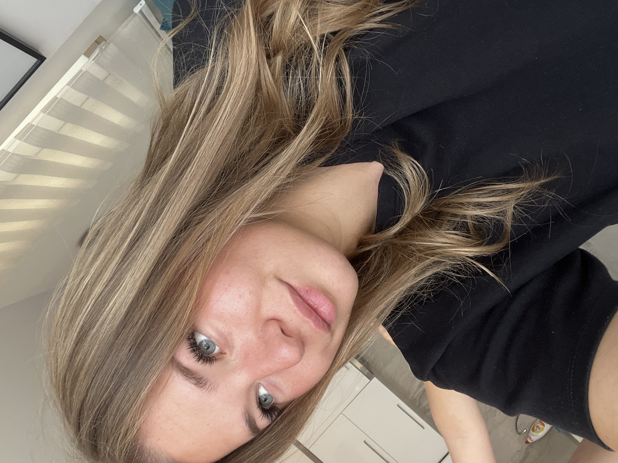 A blonde-haired woman is shown in a medium-range, slightly angled selfie. She is wearing a black t-shirt. A window with white blinds is in the upper-left corner of the frame. A kitchen counter and white cabinets are visible in the background. The location is unknown and there is no way to say it is in Budva, Montenegro.
