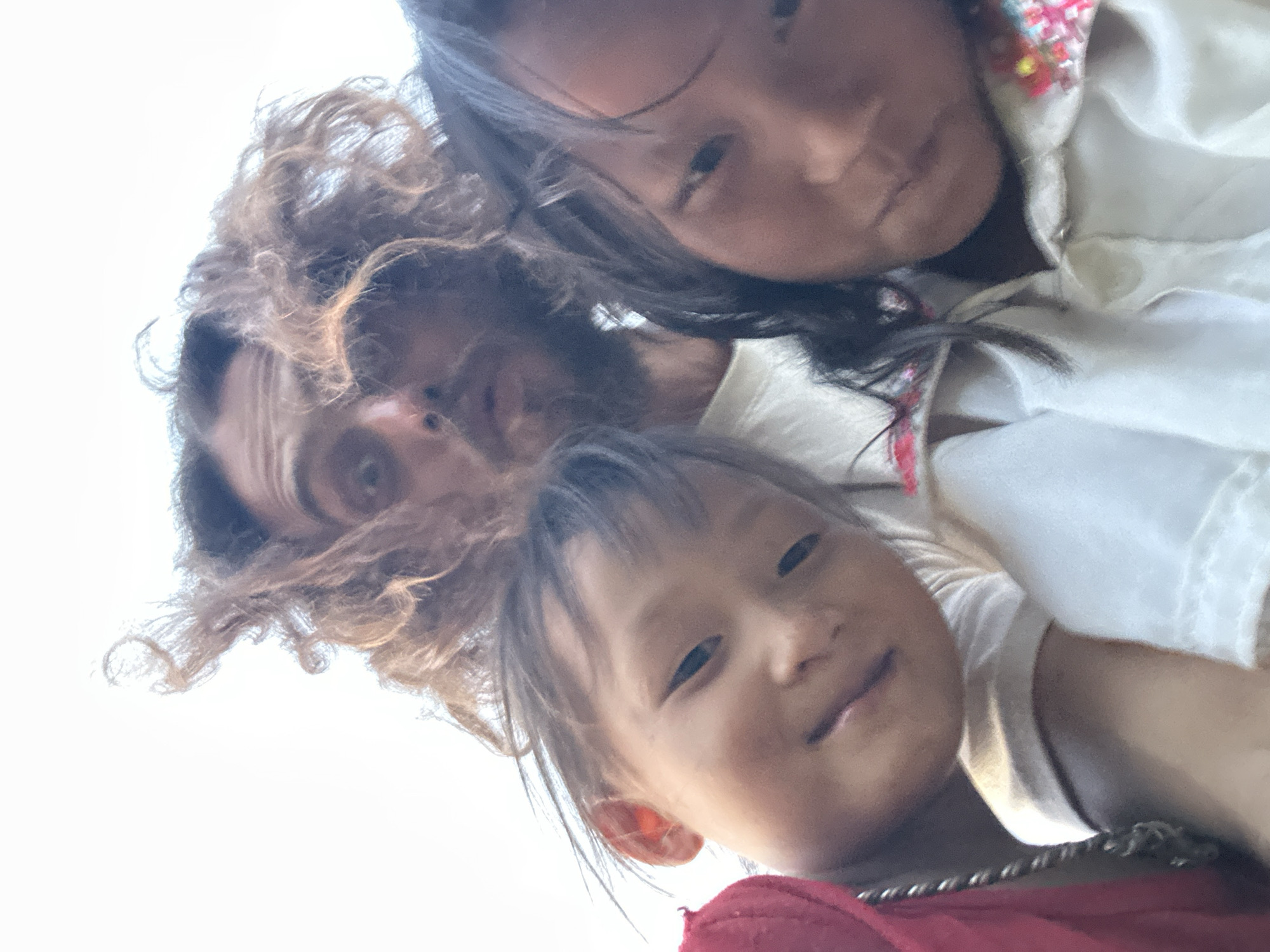 A low-angle, upward-facing photograph captures three individuals—one adult male and two children—looking down towards the camera lens against a completely overexposed white sky. The bright background suggests the image was taken outdoors during clear daylight, possibly midday or afternoon.
The adult male, positioned centrally, has curly brown hair and a beard, and looks directly into the camera with wide, curious eyes. To his upper right, one child with dark hair wears a light-colored garment featuring colorful embroidery or beadwork around the neckline, showing a neutral to slightly soft expression. Below the adult, another child with dark hair is visible, wearing a red top and a necklace, offering a gentle smile.
All subjects appear to be of Asian descent, consistent with the provided location context of Mù Cang Chải, Vietnam. The children's traditional-looking attire, particularly the embroidery, subtly hints at the cultural setting. The overall mood is informal and candid, suggesting a close-up selfie or a spontaneous moment. No discernible text is visible within the image.