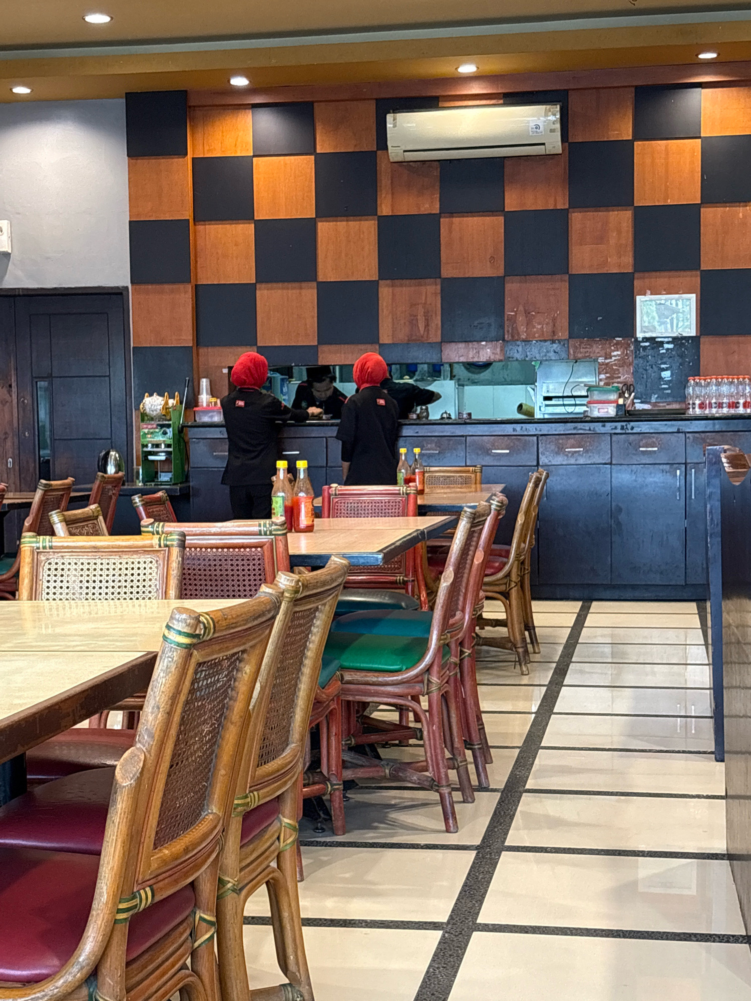 This image depicts the interior of a restaurant located in the city of Lhokseumawe, Indonesia. In the background, two individuals wearing red headscarves and black uniforms are visible behind a counter, appearing to be engaged in food preparation or service. The restaurant's wall features a distinctive checkerboard pattern of dark brown and lighter brown squares. Above the counter, an air conditioning unit is mounted on the wall. In the foreground and midground, tables and chairs are arranged, with several chairs made of woven rattan with dark red upholstery. The floor has a tiled pattern of light beige and dark gray lines. On a counter behind the individuals, bottles of water and condiments are visible.