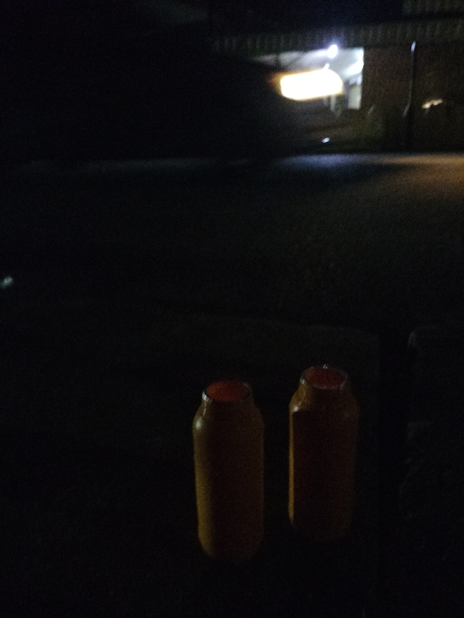 In Jalingo, Nigeria, two plastic jugs with orange lids are placed on the ground in the foreground. The jugs are illuminated by an unseen light source, casting a faint glow on their yellow bodies. The background is dark and indistinct, with a suggestion of a building or structure illuminated by a bright light, possibly indicating a commercial establishment. The ground appears to be asphalt or a similar paved surface, reflecting some ambient light. The overall scene is low-light and somewhat grainy, suggesting it was taken at night or in a dimly lit environment.