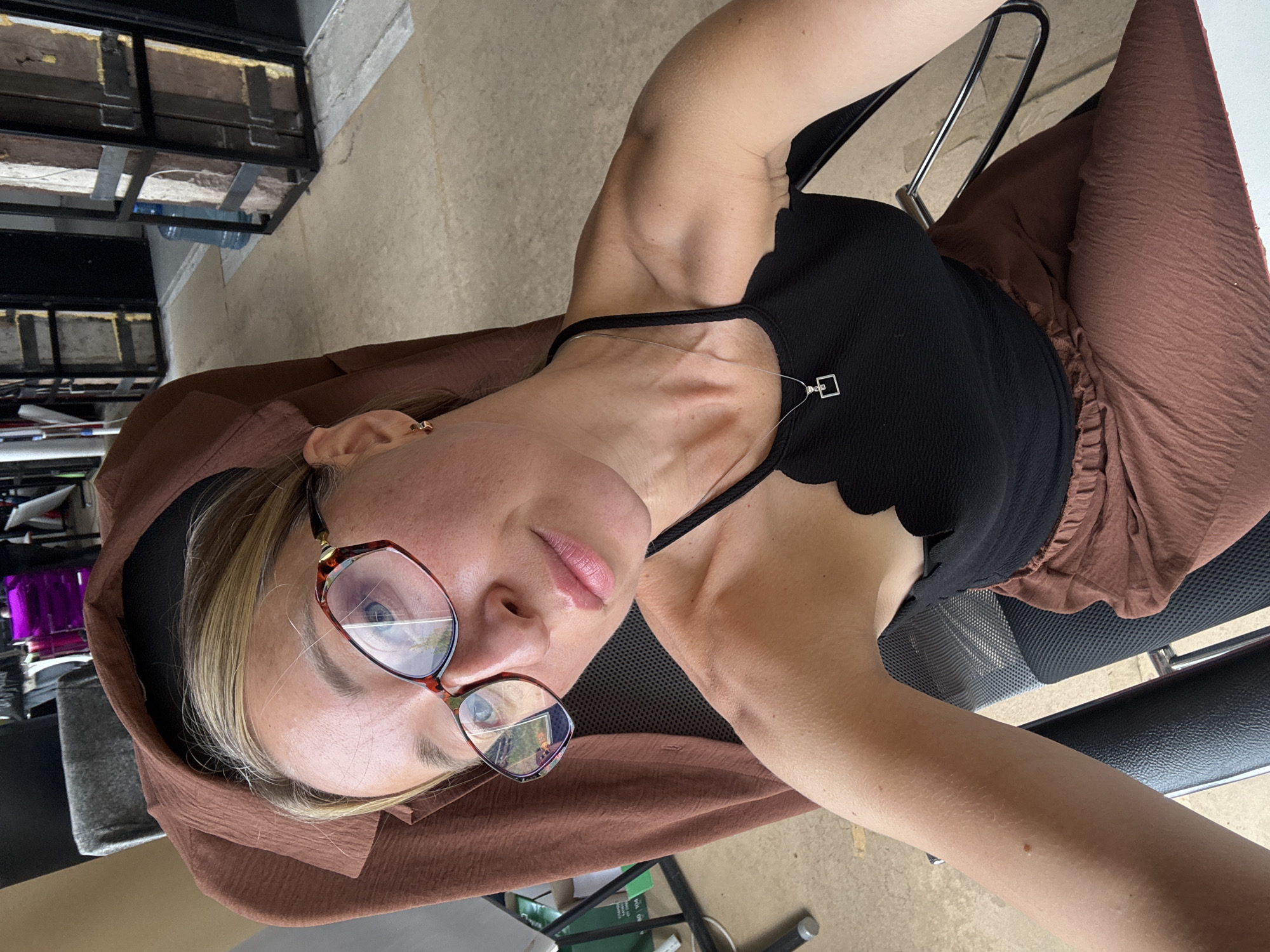 In a close-up, high-angle shot, a fair-skinned woman with blonde hair pulled back and wearing large, patterned eyeglasses is seated in an office chair. She is wearing a black halter top with a scalloped neckline and a brown, textured skirt. A silver necklace with a square pendant rests on her collarbone. Her left arm is bent and rests on the armrest of the chair, while her right arm is extended outward, likely holding the camera. The background suggests an indoor or semi-outdoor setting with concrete flooring and large, industrial-style structures. No specific location in Chaikovsky, Russia, can be identified from the provided visual information.