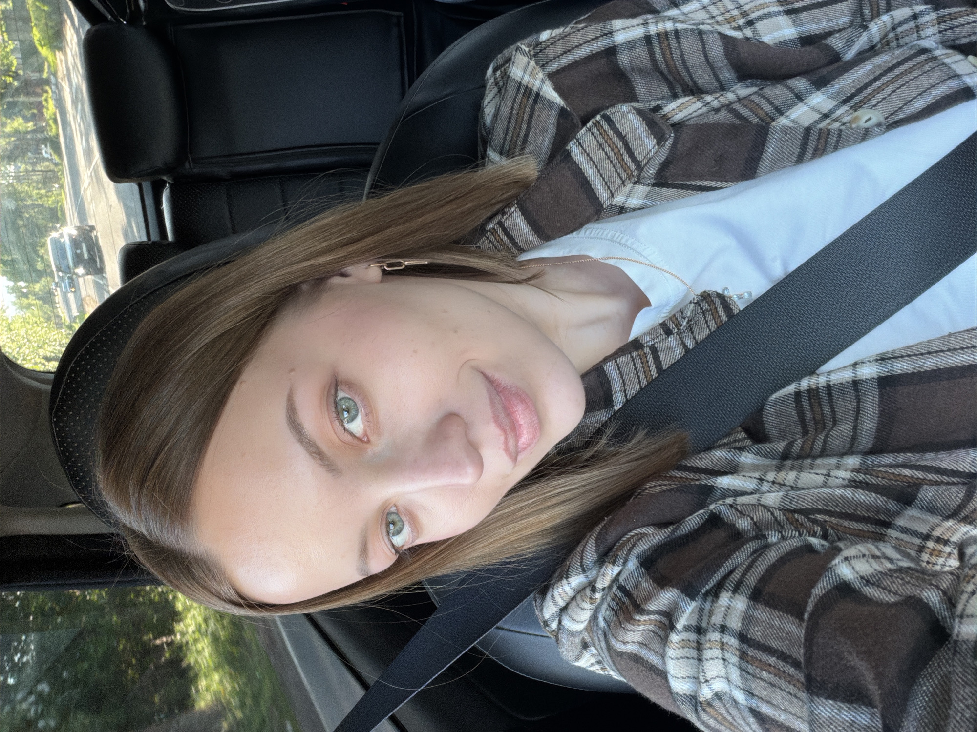 A woman with light brown hair and green eyes is in a car, wearing a white shirt and a brown plaid shirt. She is wearing a seatbelt. The location cannot be determined from the image provided. There is no indication of the city Maksimkovo, Russia.