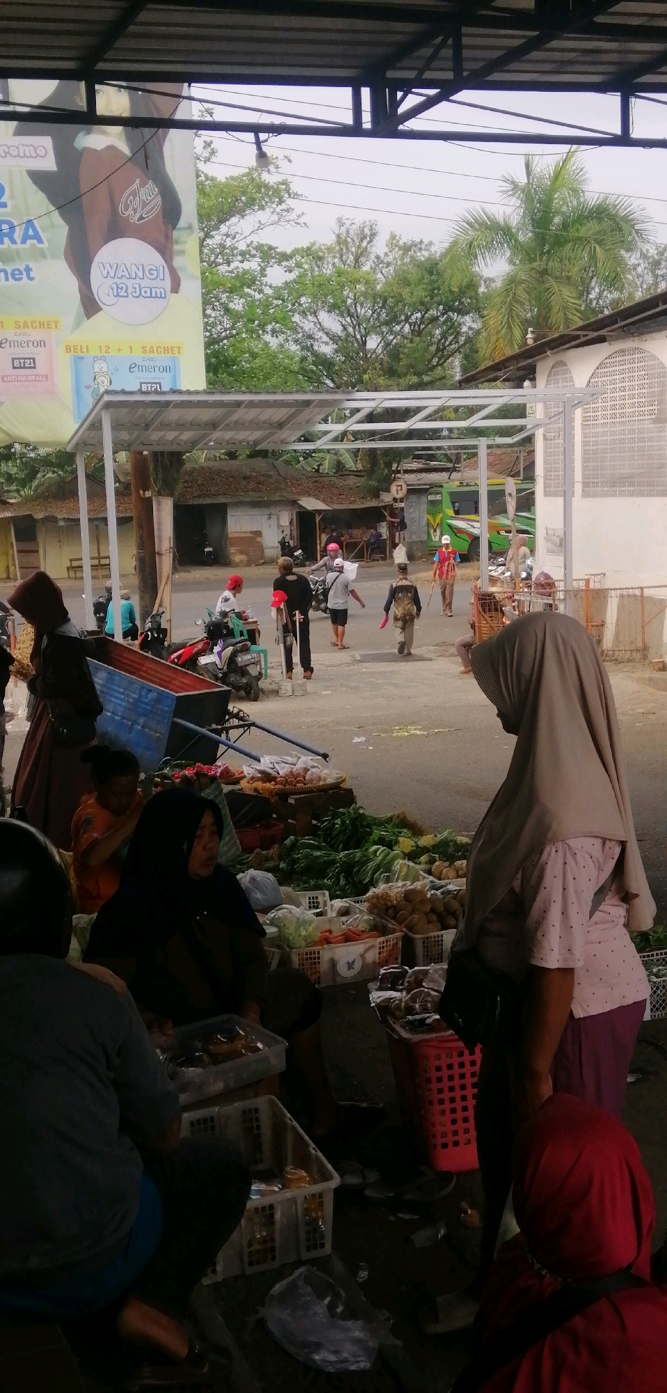 The image depicts an outdoor market scene in Ajibarang, Indonesia, likely a roadside market given the presence of vehicles and the asphalt road. The foreground is dominated by stalls laden with fresh produce, primarily vegetables like leafy greens, carrots, and possibly some fruits or other packaged food items. Several individuals are visible interacting with the market, either as vendors or shoppers.
In the immediate foreground, a person wearing a dark hijab and a light pink patterned shirt is standing and looking away from the camera, facing towards the market stalls. To the left, another person in a grey shirt and dark pants is seated, with a blue plastic bin in front of them. Further back, a woman in an orange shirt is attending to the produce.
In the mid-ground, several people are walking along the road or near the stalls. One individual in a red head covering is standing near a cart. Another person with a backpack is walking away from the camera, carrying what appears to be a red bag or cloth. A green truck or bus is visible in the background, suggesting this is a busy thoroughfare.
The overall environment appears to be somewhat overcast, with a covered market structure extending over some of the stalls. The architecture of a white building with arched windows is visible on the right side of the image, suggesting the market is situated within a developed area of the city. The presence of motorcycles parked on the side of the road indicates typical mode of transportation in the region.