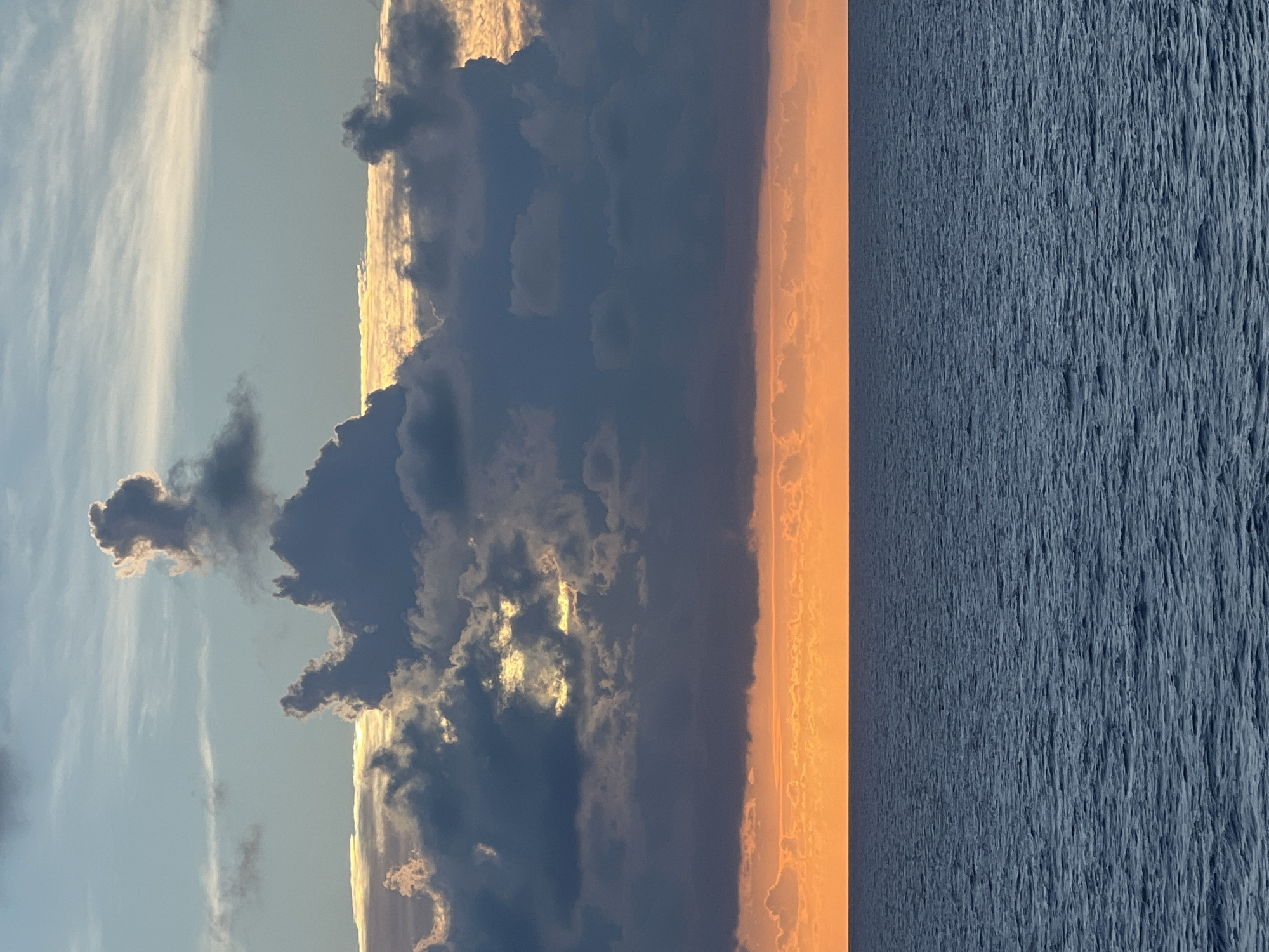 An image of the ocean at sunset is shown, with a clear horizon dividing the water and the sky. The ocean is a deep blue with gentle waves. The sky above is filled with dramatic clouds, colored in shades of gray, white, and orange. The bottom portion of the sky is a vibrant orange, indicative of the setting sun. Above the orange band, dark gray clouds are clustered, with patches of sunlight breaking through. The upper part of the sky transitions to a lighter blue with wispy white clouds. There are no people or objects visible in the scene.
