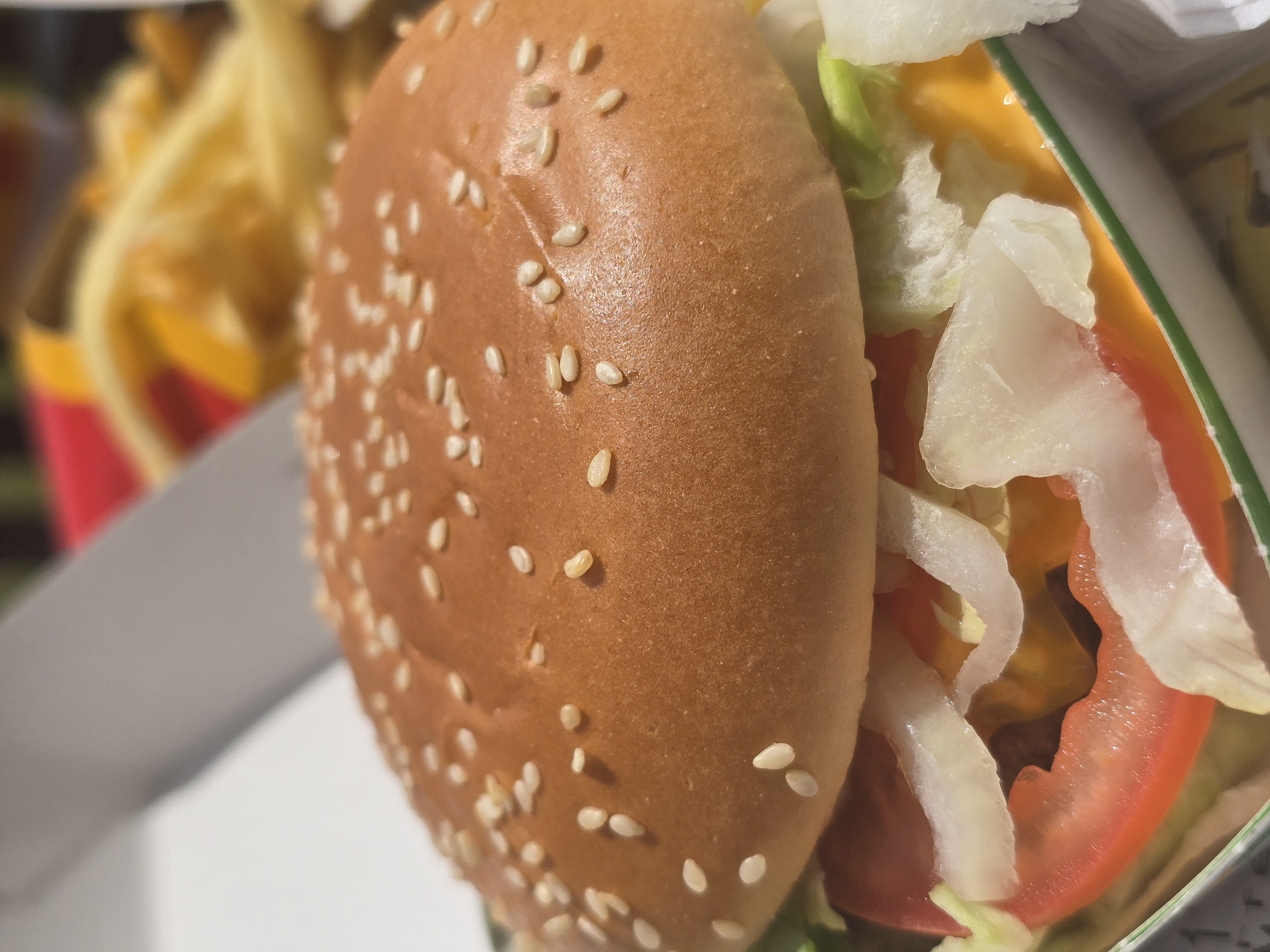 This image depicts a fast-food meal, located in Erding, Germany. The primary subject is a hamburger, centrally positioned in its open packaging, viewed from a slightly elevated angle.
The hamburger features a prominent, golden-brown top bun covered densely with white sesame seeds, exhibiting a slightly reflective surface. Beneath the bun, visible ingredients include pale green shredded lettuce, several translucent white slices or pieces of onion, and a thick, vibrant red slice of tomato. Below these, a melted, bright yellow-orange slice of cheese is visible, partially covering a dark brown meat patty. The hamburger rests within a white and green striped cardboard or paper container.
In the blurred background to the upper right, a red and yellow cardboard container, characteristic of a fast-food establishment, holds numerous out-of-focus golden-yellow french fries. The upper left background shows blurred white and light gray elements, likely part of additional packaging or a table surface. No individuals are present in the scene, and no explicit actions are depicted beyond the presentation of the meal. The scene is well-lit, suggesting either natural light or bright artificial illumination.