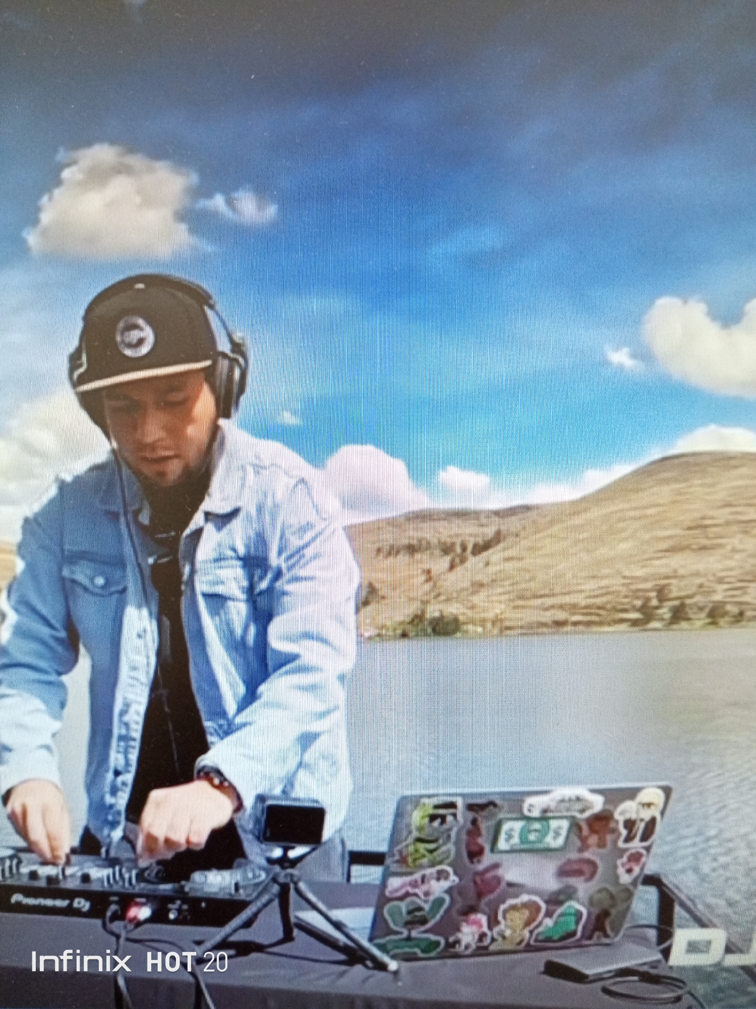 A man wearing a black baseball cap, headphones, a black t-shirt, and a denim jacket is standing in front of DJ equipment, including a mixer and a laptop covered with stickers. The man appears to be operating the mixer. The equipment is set up on a table covered with a black cloth. Behind the man is a lake and a hillside, with a blue sky and scattered clouds in the background. The camera that took the photo is an Infinix HOT 20. The location may be in the Pampatar, Venezuela area, given the described landscape features.