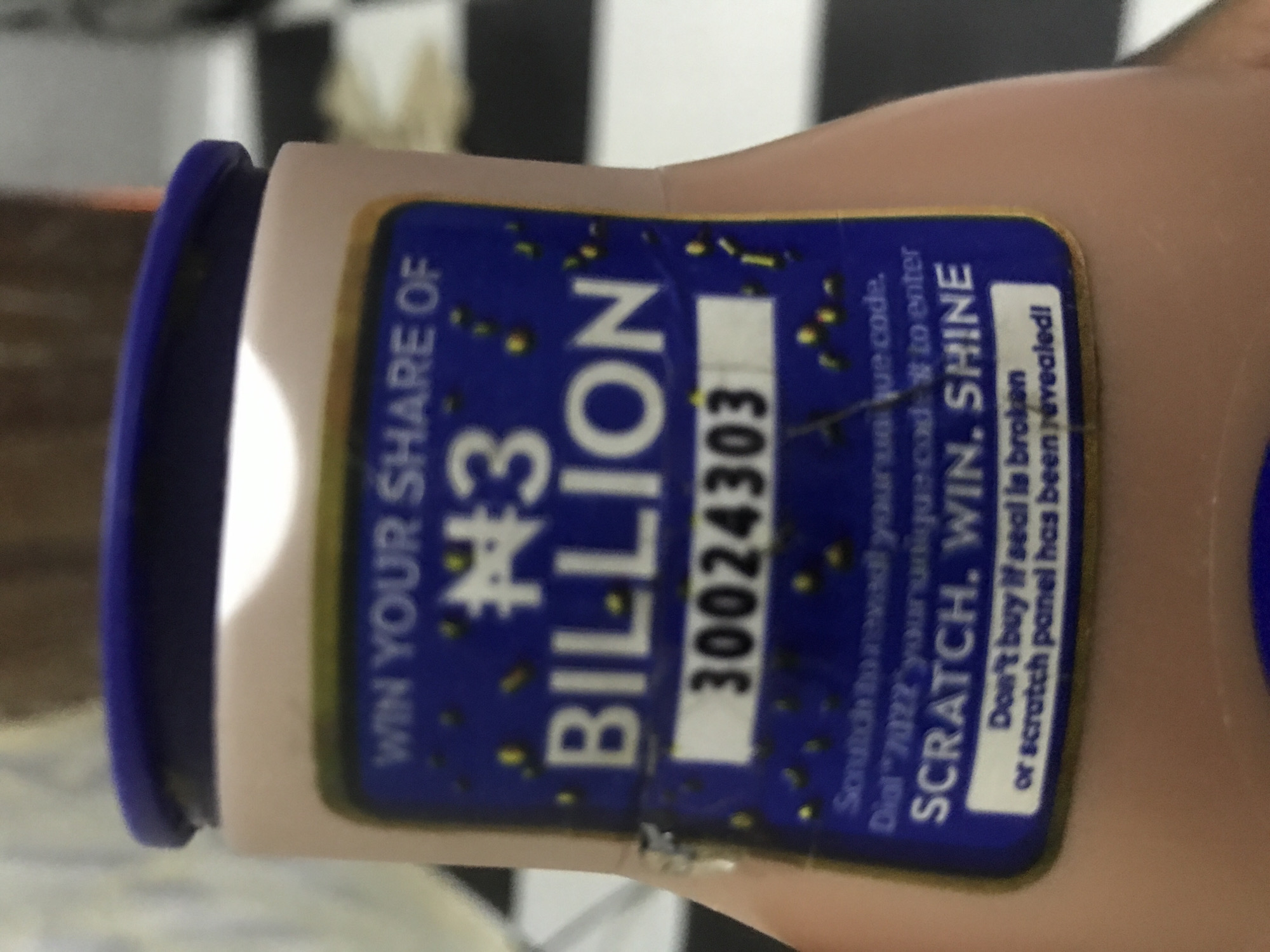 A close-up, slightly angled photograph displays a light pink/beige cylindrical product bottle, featuring a dark blue screw-top cap. Affixed to the bottle is a rectangular blue promotional label with white and yellow text. The upper portion of the label states "WIN YOUR SHARE OF N3 BILLION," where 'N' represents the Nigerian Naira currency symbol. Below this, a white rectangular panel prominently displays the numerical code "30024303." Further text, partially obscured, reads "Scratch to reveal your unique code. Dial *7022* your unique code # to enter." The bottom section of the label features the slogan "SCRATCH. WIN. SHINE" and a warning: "Don't buy if seal is broken or scratch panel has been revealed!" The blue background of the label is embellished with small yellow and dark blue speckles. The label exhibits minor wear, with a small piece of foreign material or debris adhering to its left edge. The background is out of focus, revealing a distinct black and white checkered pattern, likely a tiled floor or surface. The scene is illuminated, casting a highlight on the top of the bottle's cap and the label. No individuals are present in the image.