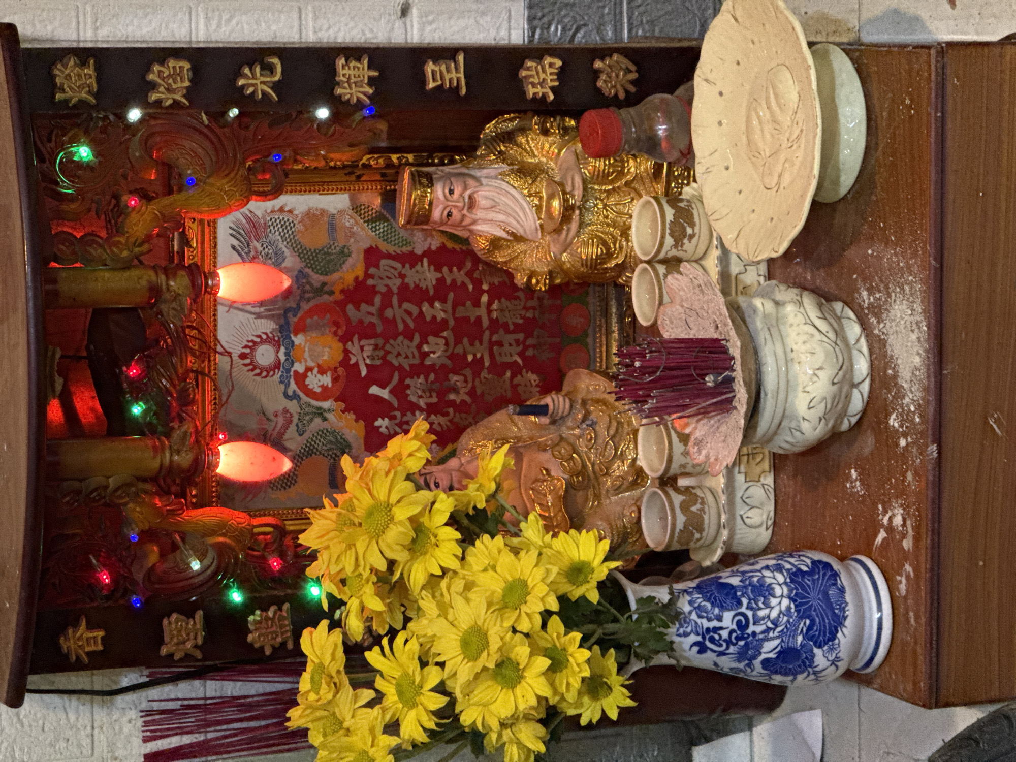 An intricately decorated domestic altar, likely located in Bac Nha Trang Ward, Vietnam, is the central focus of the image. The altar, which appears to be a wooden structure, is adorned with various items suggesting veneration and offerings.
Two prominent golden statues are placed on the altar. The one on the left, depicted with a benevolent face and white beard, is characteristic of Ông Địa (God of Earth). To its right, a statue with a longer white beard and an imperial hat represents Thần Tài (God of Wealth). Both deities are surrounded by offerings, including several small white cups, possibly for tea or water, and a red plastic-capped bottle.
In front of the deities, a cluster of yellow chrysanthemums is arranged in a white vase with blue floral patterns. Numerous thin, reddish-brown incense sticks are burning or recently burned, emitting a faint haze. A decorative, light-colored ceramic plate rests partially on the altar.
The altar is richly decorated with a red banner or scroll featuring golden text behind the statues. The surrounding wooden frame is embellished with intricate carvings and an array of colorful LED lights (red, green, blue) and two glowing red lights, illuminating the setup. The background consists of a white brick wall and a wooden surface to the right.
The visible text on the altar's main banner and surrounding plaques, as captured by OCR, includes:
- "燈焰光輝呈瑞彩" (Lantern flames radiate auspicious colors)
- "香煙盤繞五方五土龍村" (Incense smoke encircles the five directions, five lands, Dragon Village)
- "物華天宇" (Magnificence of the universe)
- "前後地主財神" (Front and back Earth Lords and God of Wealth)
- "生人傑地靈時" (When heroes are born, the land is sacred)
Other characters like "祿" (prosperity), "喜" (happiness), "壽" (longevity), "財" (wealth) are also visible on the decorative panel above.
The overall scene suggests a devotional practice, likely at night or indoors with artificial lighting, emphasizing reverence for prosperity and protection.