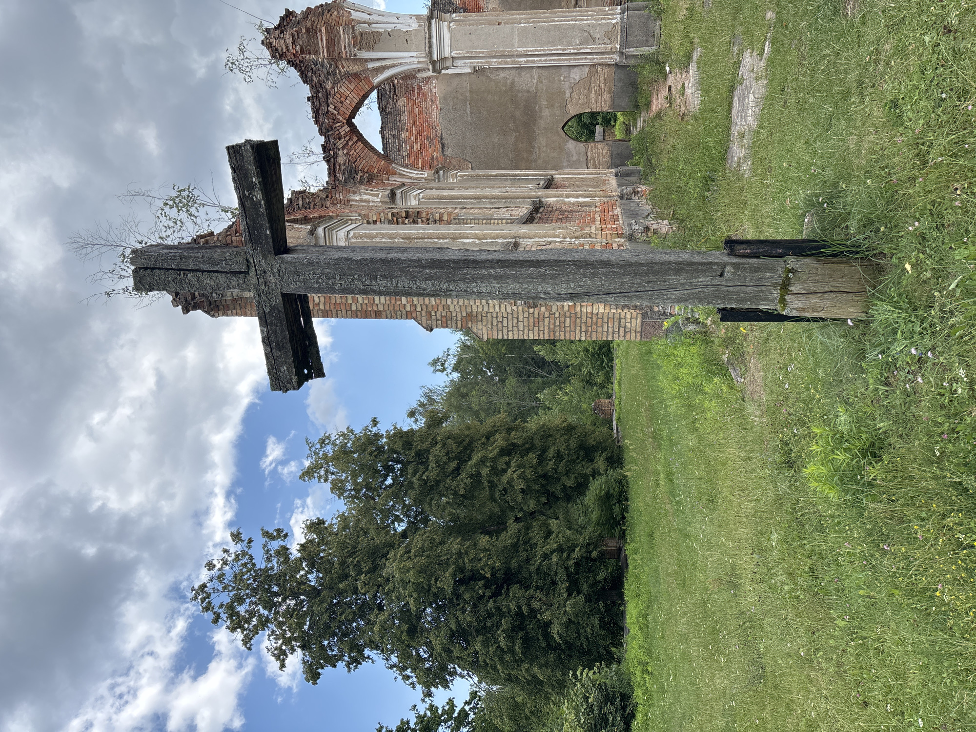 The image shows a wooden cross in the foreground, with the ruins of a brick building behind it. The building appears to be an old church, with arched windows and weathered brickwork. Trees and greenery surround the ruins, suggesting an outdoor, possibly rural, location. The sky is cloudy. The scene is set in the city of Bachury, Poland. There are no people visible in the image.