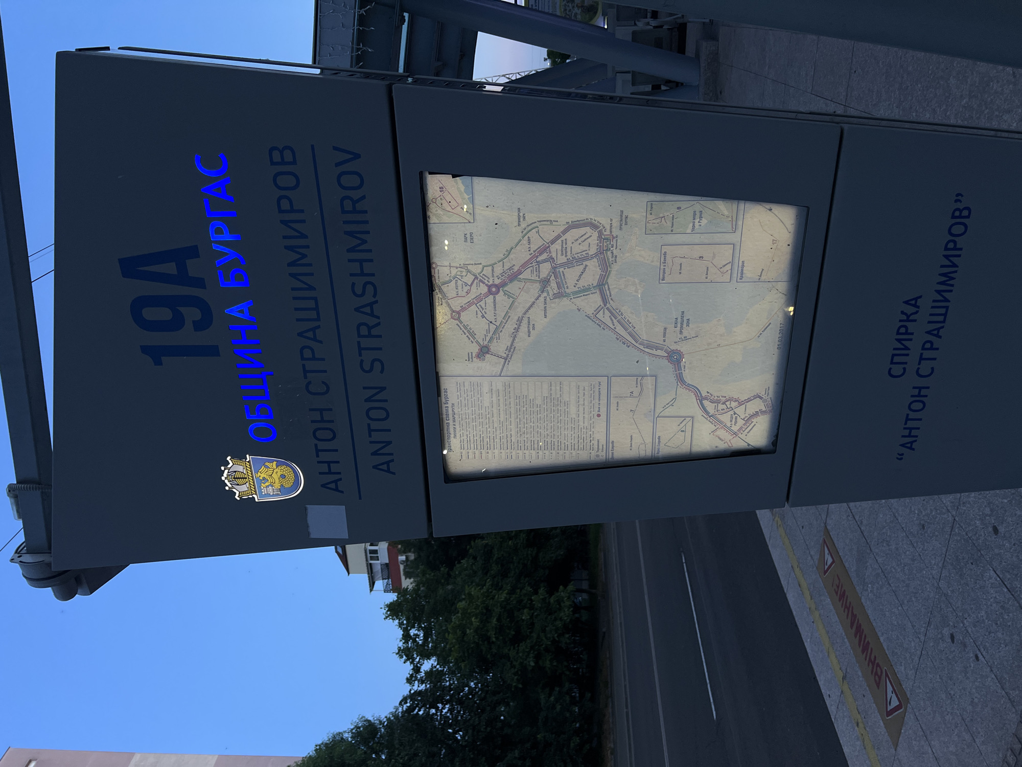 The image shows a bus stop in Burgas, Bulgaria. The bus stop has a digital display board showing a map of Burgas with bus routes. The bus stop is labeled "19A ОБЩИНА БУРГАС" and "ANTON STRASHIMIROV". The bus stop is located on a street with trees and buildings in the background. The sky is blue and clear. There is a yellow warning sign on the sidewalk with the word "ВНИМАНИЕ" in red letters. The sign indicates caution.