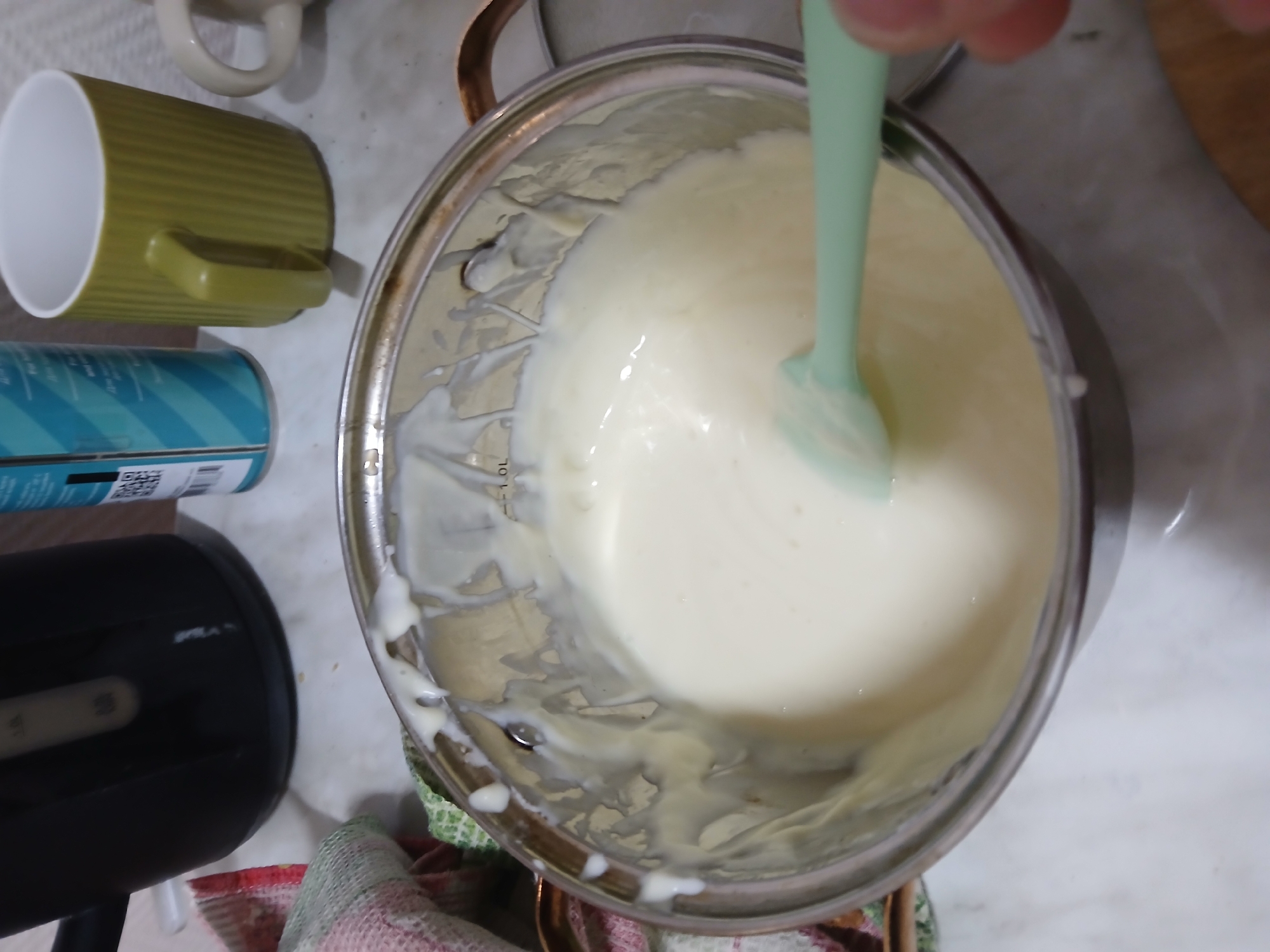 An overhead shot shows a kitchen scene in Chaikovsky, Russia. A stainless steel pot filled with a thick, white liquid is the central focus. A light green spatula stirs the substance. Adjacent to the pot is a green mug, a blue aerosol can, and a black appliance, potentially a mixer. A red and green cloth lies partially under the pot. The background consists of a white countertop.