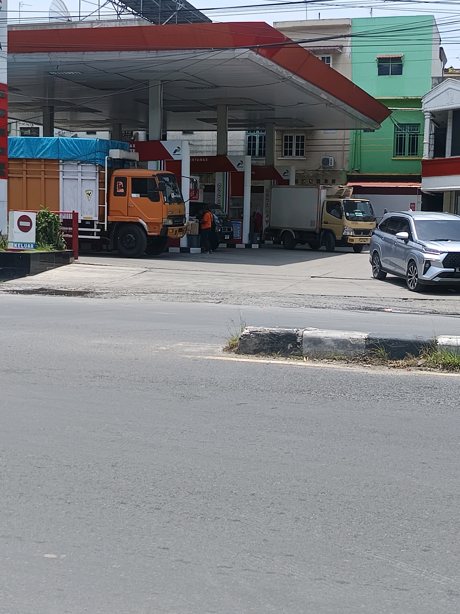 In Percut Sei Tuan, Indonesia, a bright, sunny day is captured at a gas station. The scene features multiple vehicles, including an orange truck with a blue tarp covering its cargo, a white delivery truck, and a silver SUV. A person in an orange shirt is visible near the orange truck, and another individual in darker clothing stands by the white truck. Behind the gas station, a light green and beige building with windows is visible, along with a white building with arches. The foreground shows a road with a concrete divider and some sparse vegetation. The image provides a snapshot of daily activity at this location.