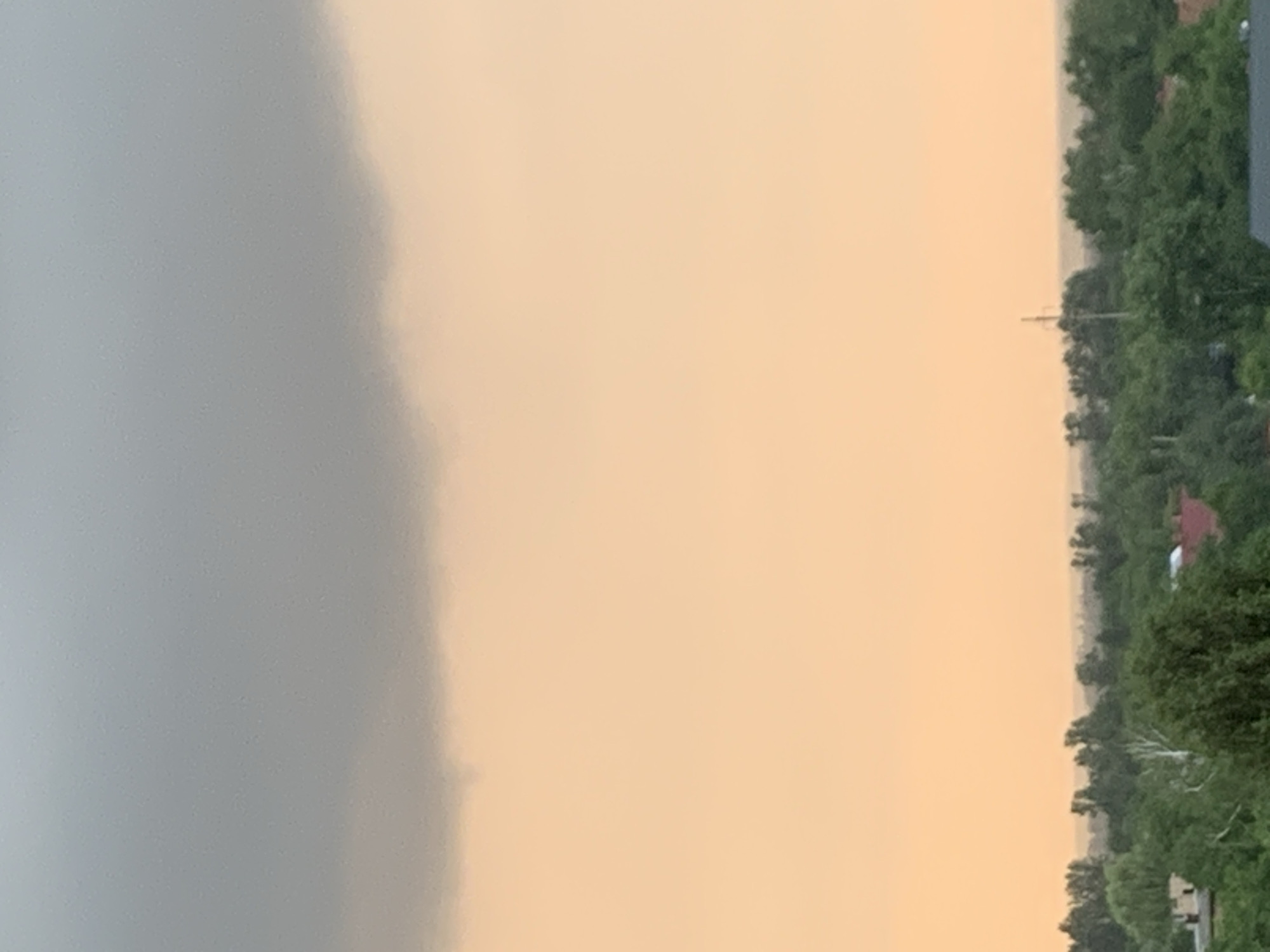 The image shows a landscape view with a heavy, dark cloud formation dominating the upper portion of the sky. Below the cloud, the sky is illuminated with a peachy, orange glow, suggesting either sunset or sunrise. The lower section of the image displays a dense canopy of green trees, indicating a heavily wooded area. Some rooftops of buildings are visible peeking through the foliage, suggesting a populated area or a town interspersed with natural greenery. The horizon is hazy, obscuring distant details. The overall impression is of a dramatic sky over a lush, possibly rural or suburban, environment. This scene is located in Kolomyia, Ukraine.