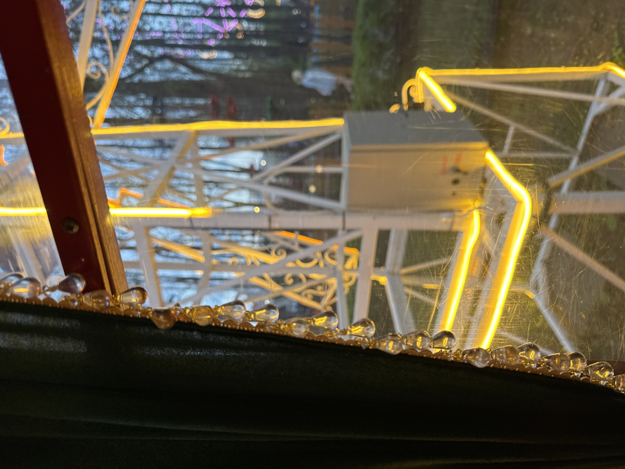The image shows a close-up, partially obstructed view, likely taken from inside an enclosed structure. The foreground features dark green fabric adorned with a string of small, clear, droplet-shaped beads. Beyond this, the dominant visual elements are components of a Ferris wheel or similar amusement ride, constructed of white metal and illuminated with warm yellow lights. The structure has intricate decorative details. Through the blurred background, vague shapes suggesting trees, foliage, and distant purple lighting can be discerned. The presence of an amusement ride with decorative lighting implies the setting is likely a park, fairground, or entertainment area. The reference to Perm, Russia, suggests this is where the image was taken, though this is not explicitly visible in the frame. There are no people visibly interacting in the image.