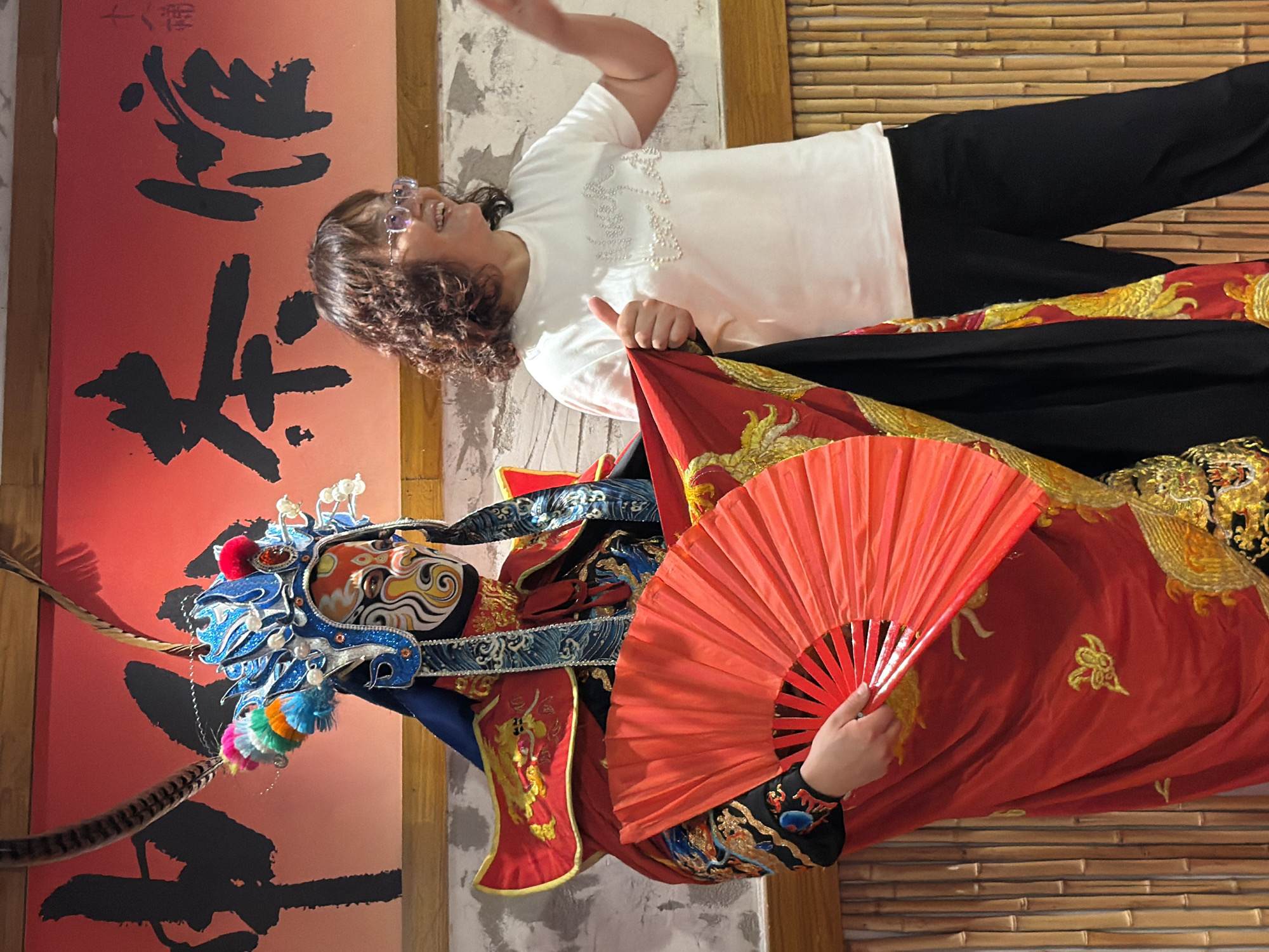 A woman in a white t-shirt and black pants stands to the right of a performer dressed in a traditional Chinese opera costume. The performer wears an elaborate blue and gold headdress with feathers and a colorful mask, and holds a red fan. The background features a red banner with black calligraphy and a wall of bamboo. The scene appears to be indoors, possibly in Yuzhong District, China, given the cultural context. The woman is smiling and giving a thumbs-up gesture, interacting with the performer.