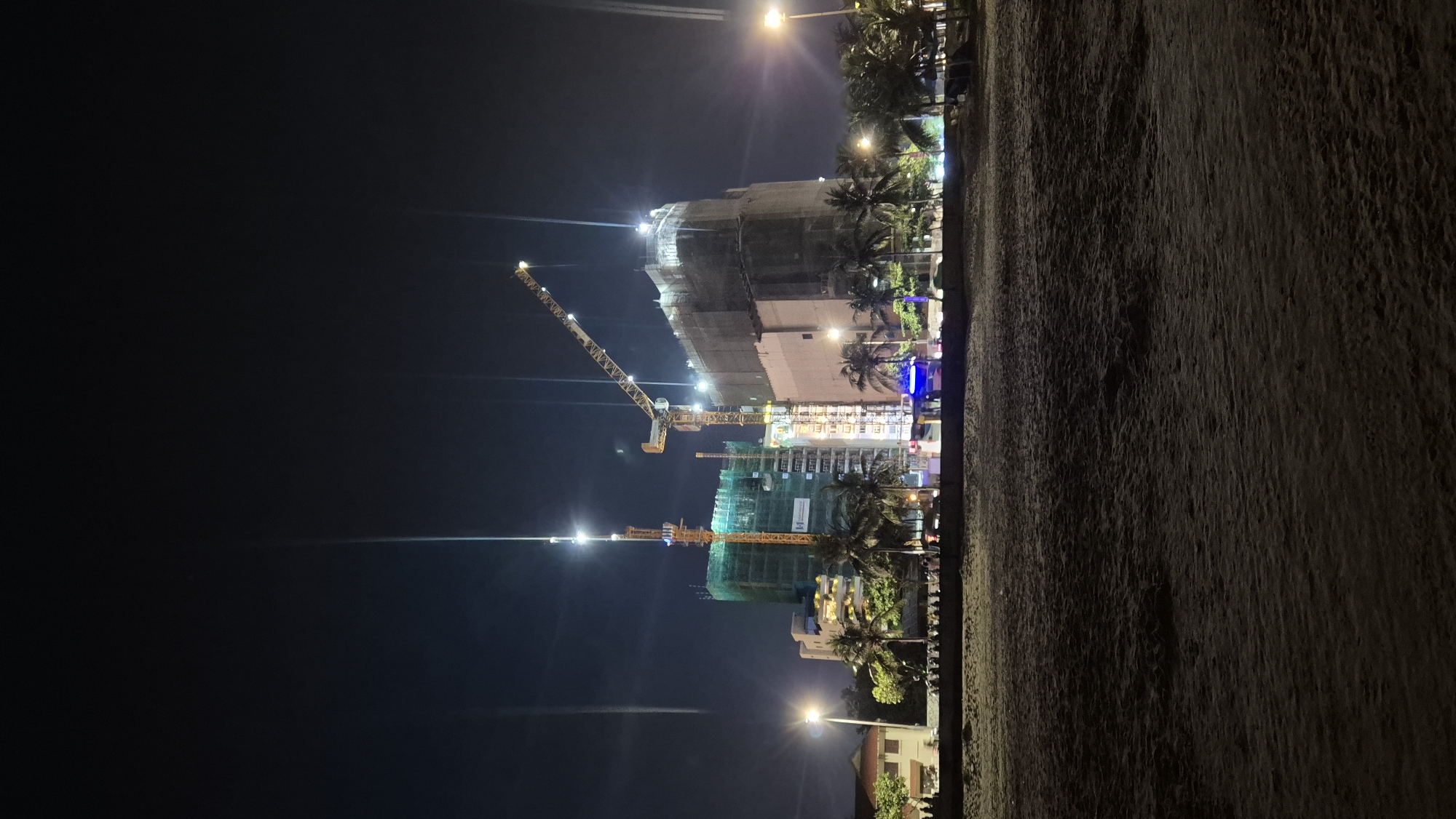 This image depicts a nighttime scene at a construction site in Da Nang, Vietnam, likely near the beach given the presence of sand in the foreground and palm trees. Two large construction cranes are prominent, one extending over a tall building covered in scaffolding and netting, and another taller crane positioned to the left. The buildings are illuminated by artificial lights, highlighting the ongoing construction. In the middle ground, a line of palm trees separates the construction site from the beach. A low wall or barrier runs along the edge of the beach, behind which a group of people are gathered, appearing to be observing the construction or enjoying the evening. Streetlights are visible in the distance, illuminating the area and adding to the overall nighttime ambiance. The sky is dark, indicating it is night.