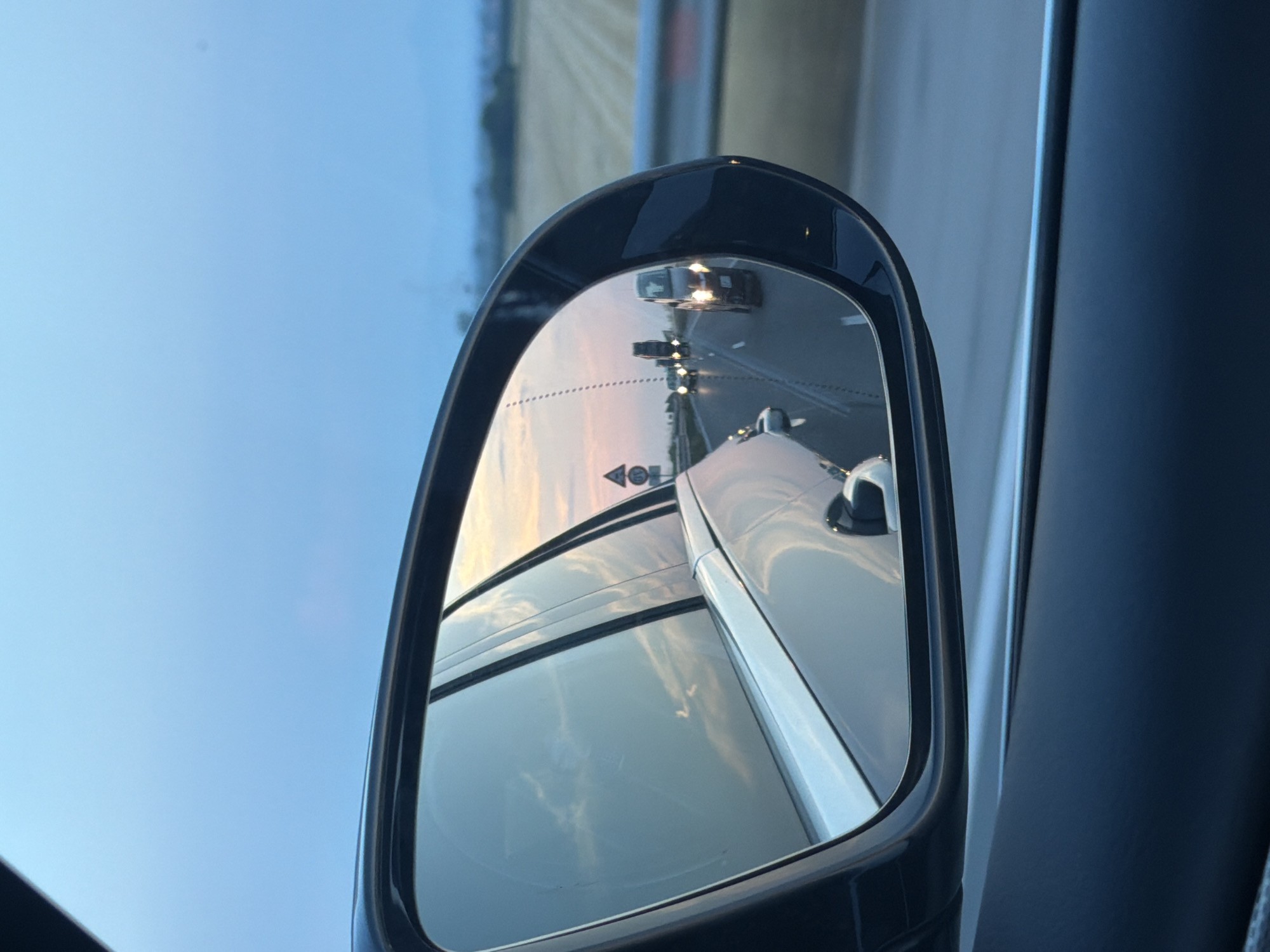 An image captured in Mirovo, Bulgaria, depicts a view from the interior of a vehicle, primarily focusing on its driver's side rear-view mirror and the external environment at what appears to be dusk. The foreground of the image features the dark trim and interior paneling of the car, with the large, dark-framed side-view mirror prominently positioned on the right.
Reflected within the mirror is a multi-lane highway, with traffic moving in the same direction as the observer. Several vehicles are visible, all with their headlights illuminated. The closest reflected vehicle, a compact car or utility vehicle, faces the observer directly, its headlights distinct. Further down the highway, additional vehicle lights are discernible. A dashed line, indicative of a blind spot monitoring system or a mirror warning area, is present on the left side of the mirror's reflection. A triangular road sign with a red border and a black symbol is also visible on the roadside within the reflection. The reflected sky above the highway displays a gradient from light blue to soft pink and orange hues, characteristic of twilight.
Beyond the mirror's frame, on the right side of the primary image, a wide, flat agricultural field with visible crop rows extends towards the horizon. A barrier or guardrail runs along the right edge of the highway. The overarching sky, visible above the car's structure and the distant landscape, is a clear, pale blue, transitioning subtly towards the horizon. The environment suggests a rural or semi-rural setting alongside a well-maintained highway. No people are explicitly visible in the scene.