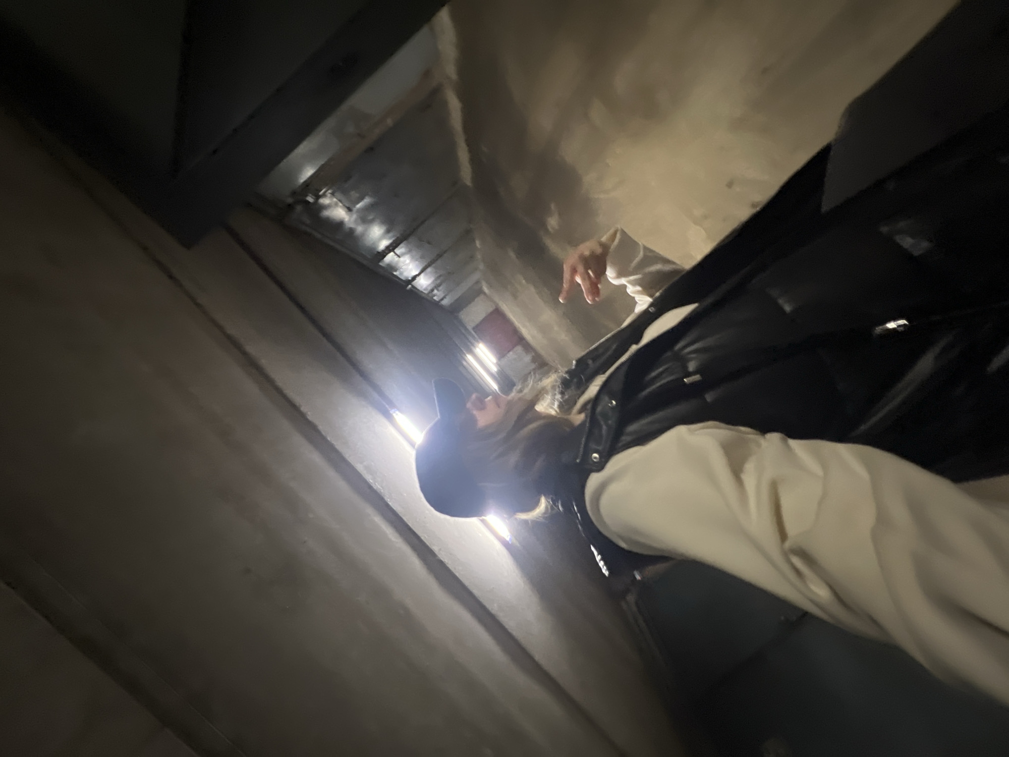 A low-angle shot depicts a person in a black cap, a black puffer vest, and light-colored pants standing in what appears to be a concrete tunnel or underpass. The walls are gray and textured, and the lighting is uneven, with a brighter area in the distance suggesting an exit. The photograph is taken from a ground-level perspective, emphasizing the height and structure of the tunnel. The location is not discernible from the visual information provided.