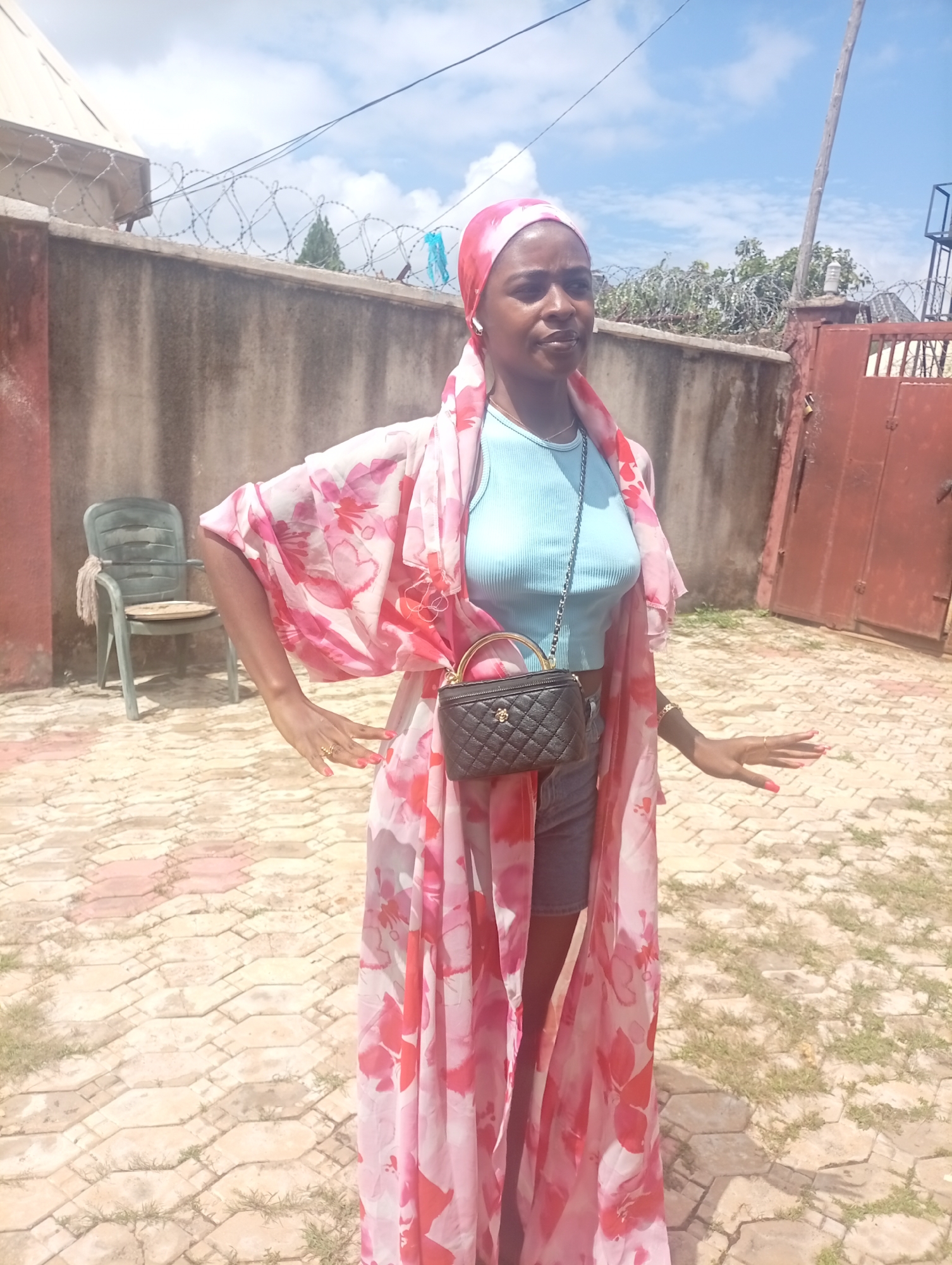 A medium-shot, eye-level, full-body photograph captures a young Black woman standing outdoors in what appears to be a residential courtyard in Rido, Nigeria. The woman is positioned in the center of the frame, facing the camera with a neutral expression and her body angled slightly to her left. She wears a light blue ribbed tank top and denim shorts, partially obscured by a long, flowing pink and red floral-patterned kimono. A small black quilted handbag with a gold chain strap hangs from her right shoulder, and she wears a gold ring on her left hand. Her head is covered with a pink and red patterned headscarf.
The background consists of a neutral-toned concrete wall with barbed wire running along its top. To the left of the woman, a green plastic chair with a worn seat is visible. To the right, a large, dark red metal gate stands ajar, revealing a glimpse of what might be a staircase structure beyond it. The ground is paved with interlocking beige and pink hexagonal tiles, interspersed with patches of grass. The sky above is a clear blue with scattered white clouds, suggesting a sunny day. The overall impression is a candid shot taken in a casual, possibly domestic, setting.
