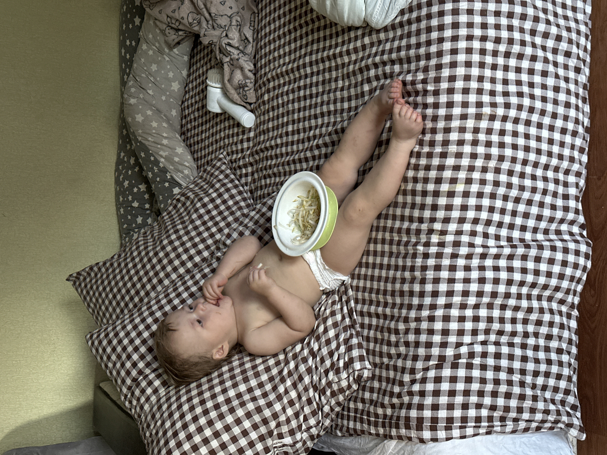A baby lies on a brown and white checkered bedspread in what appears to be a room with yellowish walls. The baby is wearing a diaper and has a bowl of sprouts or shredded vegetables resting on their stomach. They are lying on a matching checkered pillow and appear to be putting their fingers in their mouth. A white baby monitor is visible at the top of the frame near some grey patterned fabric. The scene is likely indoors, as the lighting suggests. The location is unspecified and not discernible from the image.