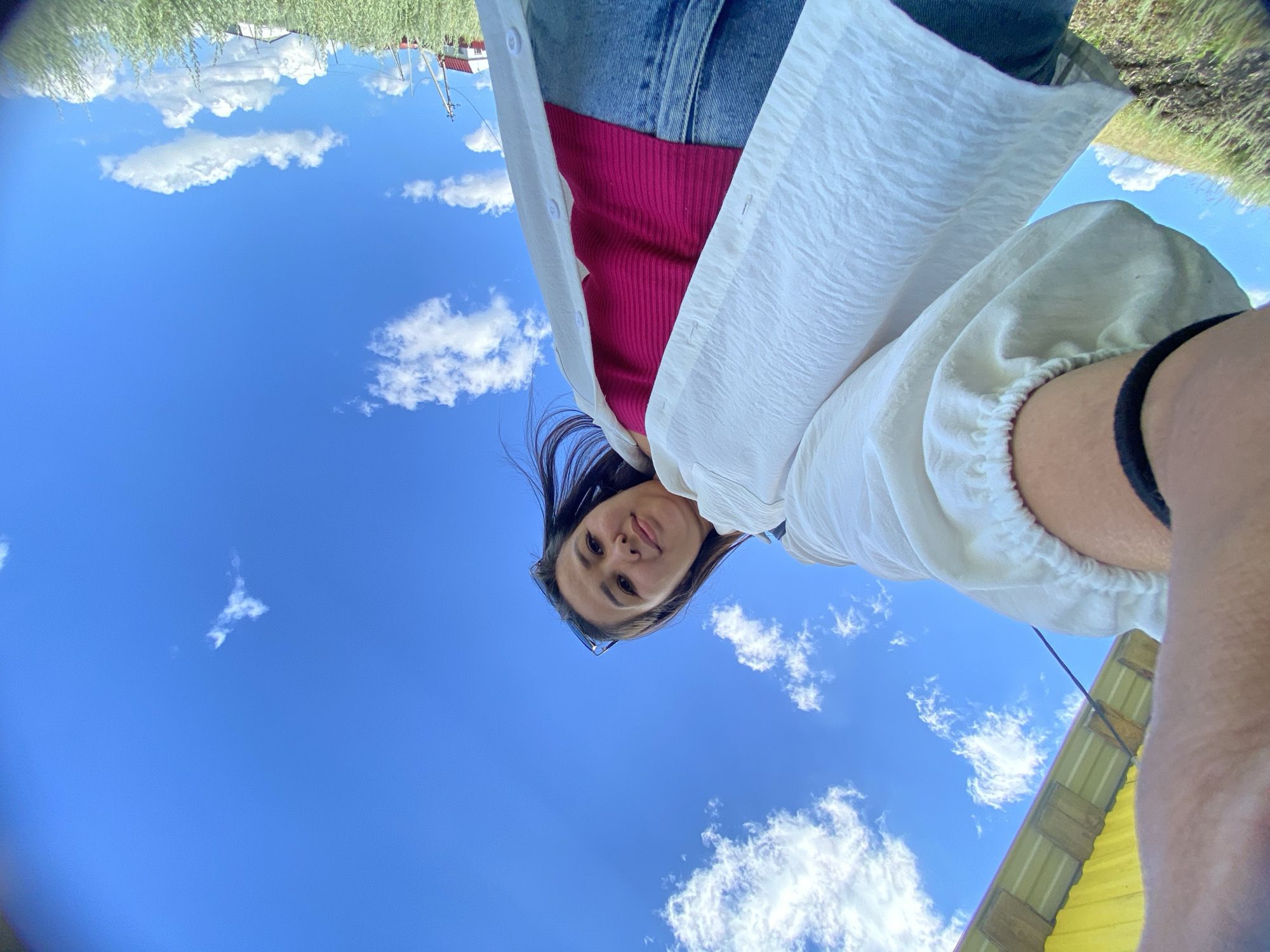 A low-angle, wide-angle shot captures a young woman looking up at the camera from below, framed against a bright blue sky with scattered white clouds. She is outdoors, possibly in Yuldybayevo, Russia, given the grassy hillside and distant structures. The woman has long, dark hair and is wearing a white textured shirt over a pink ribbed tank top and blue jeans. Her left arm is raised, holding the camera, with a black elastic band visible on her bicep. In the background, a yellow structure with visible wooden beams is partially visible at the top left of the frame. The landscape features dry, green grass and a few small buildings in the distance, with power lines visible to the right. The overall impression is one of a casual, outdoor moment.