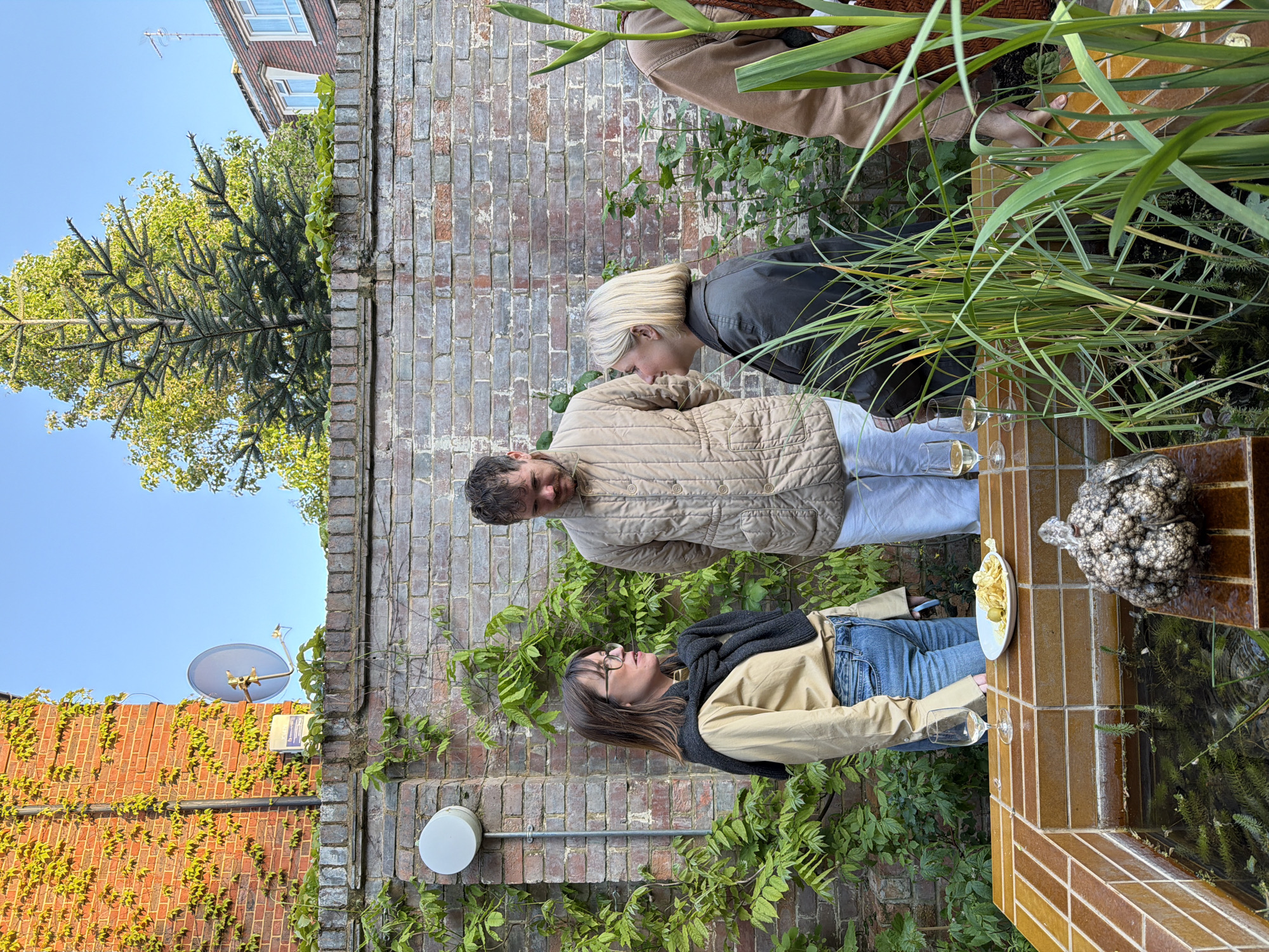 This media file captures an outdoor social gathering of three adults in what appears to be a residential garden or patio area in London, United Kingdom, during the late afternoon or early evening. The clear blue sky and long shadows suggest pleasant weather.
In the foreground, a woman with long brown hair and glasses, wearing a beige jacket, blue jeans, and a dark scarf, stands facing towards the right side of the frame. She holds a clear glass in her hand, with a plate of green olives and what look like potato chips on a tiled surface beside her. This tiled surface, possibly an outdoor bar or planter, also holds several other clear glasses, some containing a light-colored beverage, and a distinctive, textured, grayish-brown decorative object resembling a large cauliflower or coral. Water is visible within the tiled structure, reflecting light.
Behind her, a man with a beard and brown hair, dressed in a beige quilted jacket and light trousers, stands looking towards the woman. To his right, partially obscured by tall green grasses, a blonde woman with short hair and a dark jacket is leaning in, seemingly engaging in conversation with the man. Another person's arm and torso, wearing a brown top, are visible behind the blonde woman, further to the right.
The background features brick walls, some covered in ivy, creating a traditional urban garden aesthetic. Trees, including a conifer and deciduous varieties, are visible against the bright sky. A residential building with windows and a television antenna can be seen in the distance, above the brick wall. A satellite dish and a white circular outdoor light fixture are mounted on the brickwork. On one brick wall, a blue sign is partially visible with the text "BTP 020...", likely indicating a London telephone number prefix, supporting the location context. The overall atmosphere appears relaxed and convivial, indicative of an informal social event.