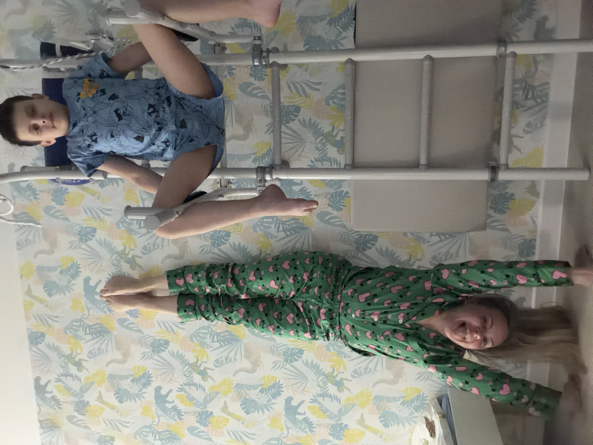 A full-body shot captures two children inside a room, likely in Votkinsk, Russia, against a wall covered with patterned wallpaper featuring light blue, yellow, and beige tropical leaves and animal silhouettes, possibly monkeys and big cats. The image is presented sideways but depicts subjects as if they are lying on their backs.
On the left side of the frame, a young boy lies on his back. He wears a light blue t-shirt with a yellow cartoon character and blue shorts. His legs are elevated and secured by white straps to a white metal frame, suggesting he might be undergoing rehabilitation or therapeutic stretching. He gazes towards the camera with a neutral expression.
To the right, a young girl lies on her back, clad in vibrant green pajamas adorned with pink hearts. Her long, light-colored hair spreads out around her head. Her arms are stretched above her head, and her bare feet are visible. She is smiling broadly at the camera, radiating happiness and playfulness.
A white metal frame, possibly part of a bed or specialized equipment, extends across the scene, dividing the space between the children and running in front of the patterned wall. The indoor setting shows no specific time of day or weather cues. There is no visible text in the image.